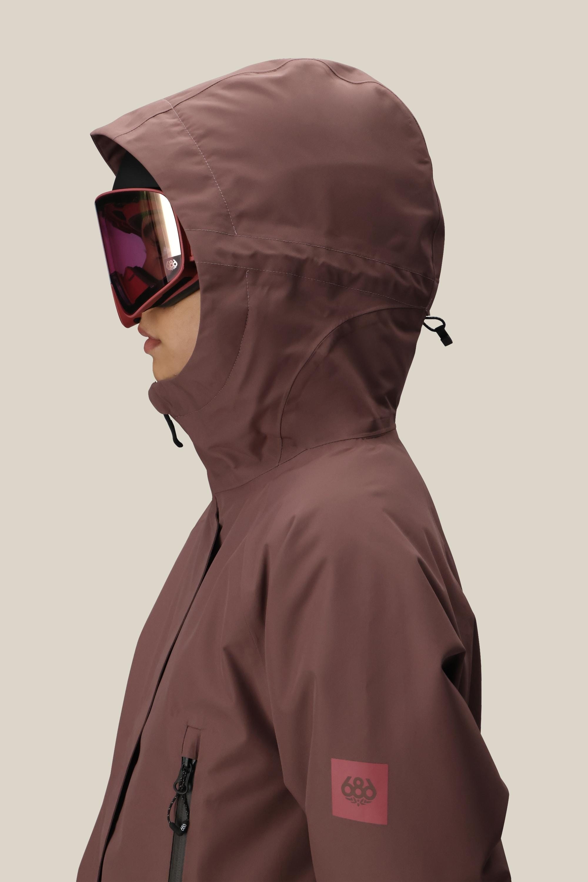 A person in profile wears the 686 Womens Whisper Insulated Jacket—a mauve, waterproof polyester jacket with a hood, zipper pocket, and sleeve patch logo—paired with reflective ski goggles, set against a plain beige background.