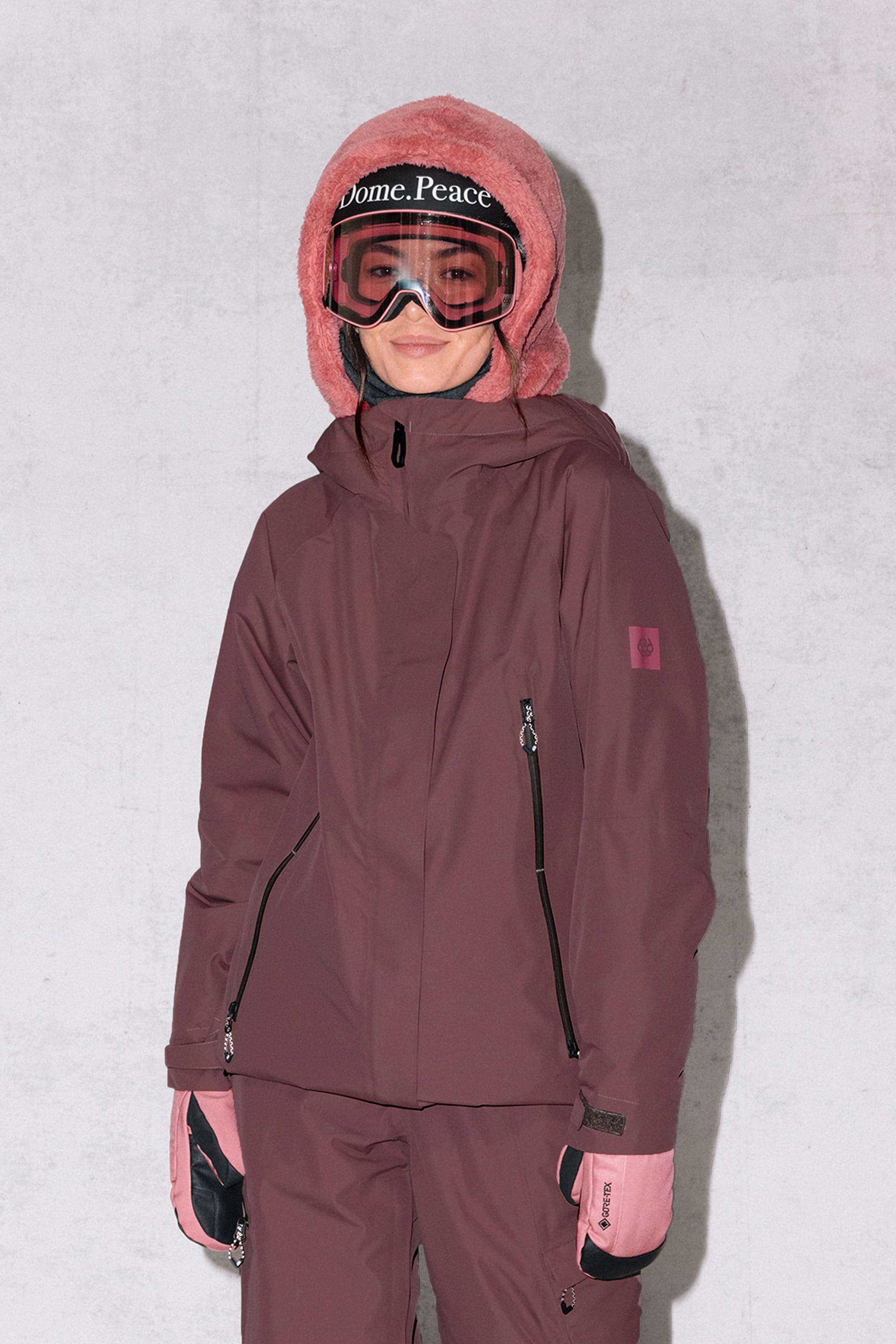 A person wearing the 686 Womens Whisper Insulated Jacket in maroon, matching pants, pink gloves and hood, and ski goggles with Dome. Peace on the strap, stands against a plain light background.