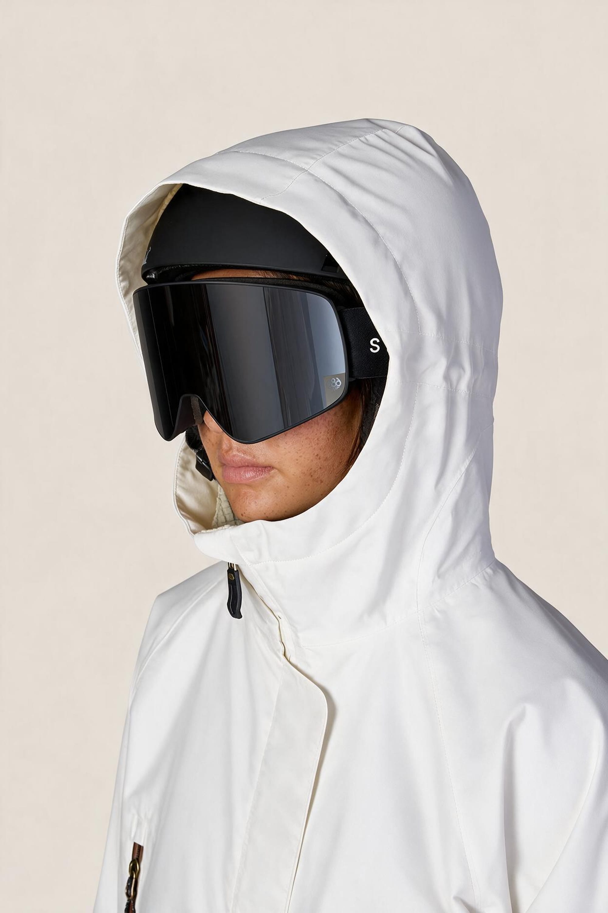A person wearing the 686 Women’s Whisper™ Insulated Jacket in white, paired with large black ski goggles and a black helmet, is facing left against a plain light background.