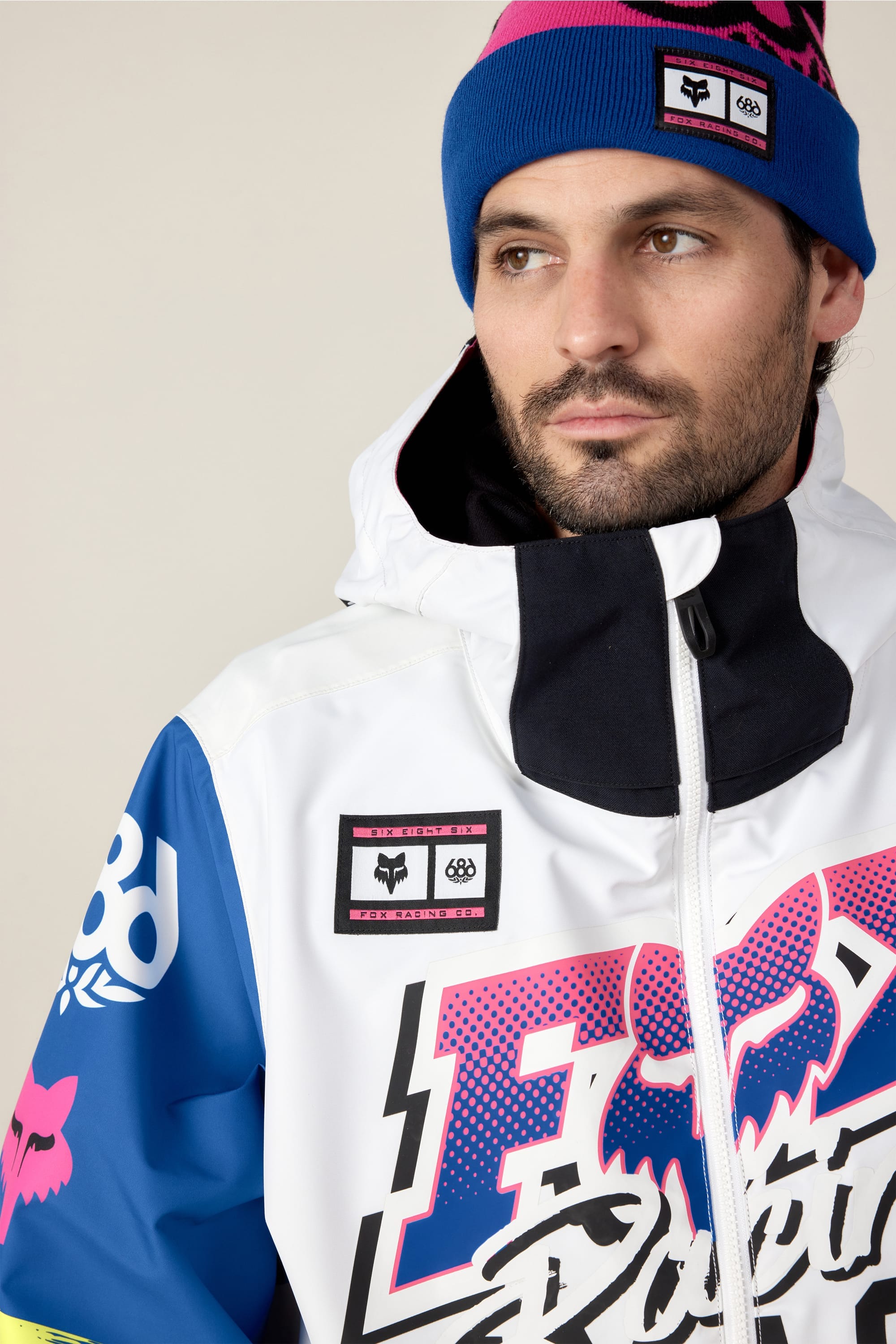 A bearded man wears the 686 Men's Fox Racing Shell Anorak, a vibrant winter jacket with bold graphics, and a blue beanie. He looks off to the side against a plain, light-colored background.