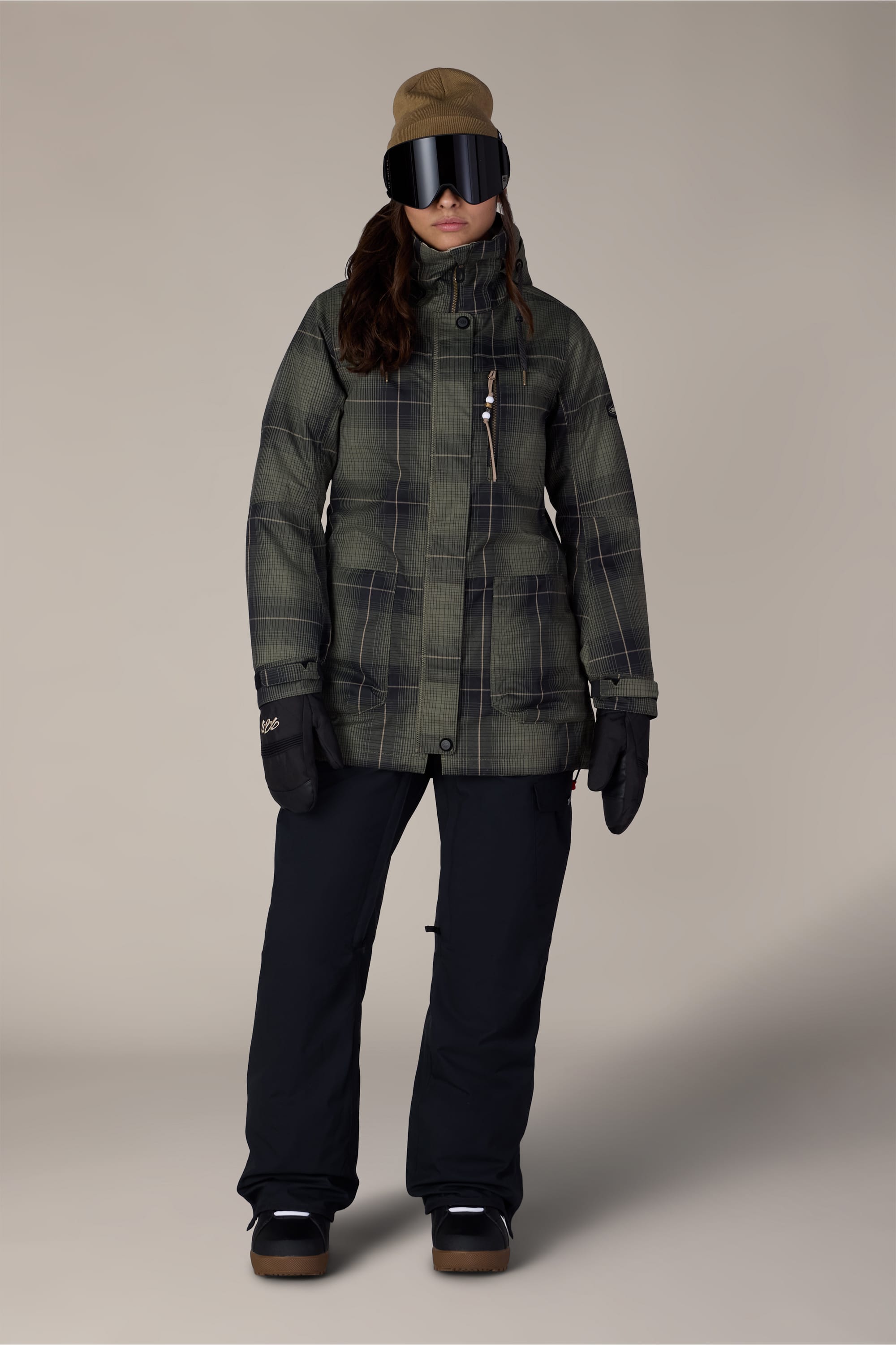 A person stands facing forward against a plain background, wearing the 686 Women’s Black Magic™ Insulated Bib by 686 paired with a green plaid winter jacket, black gloves, brown hat, and large ski goggles.