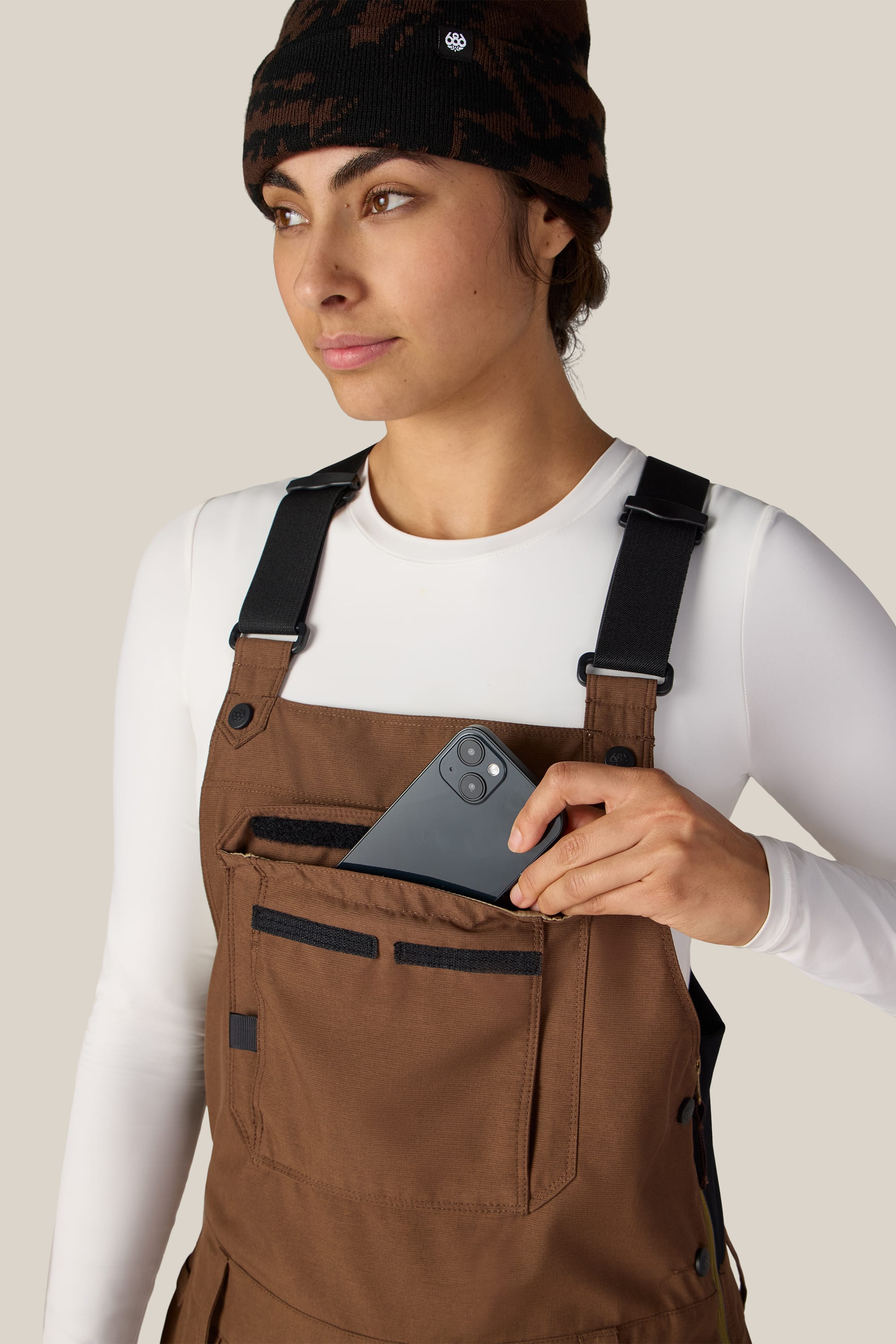 A person wearing the 686 Women’s Black Magic™ Insulated Bib by 686, along with a white long-sleeve shirt and a patterned beanie, puts a smartphone into the front pocket of their bib against a plain background.