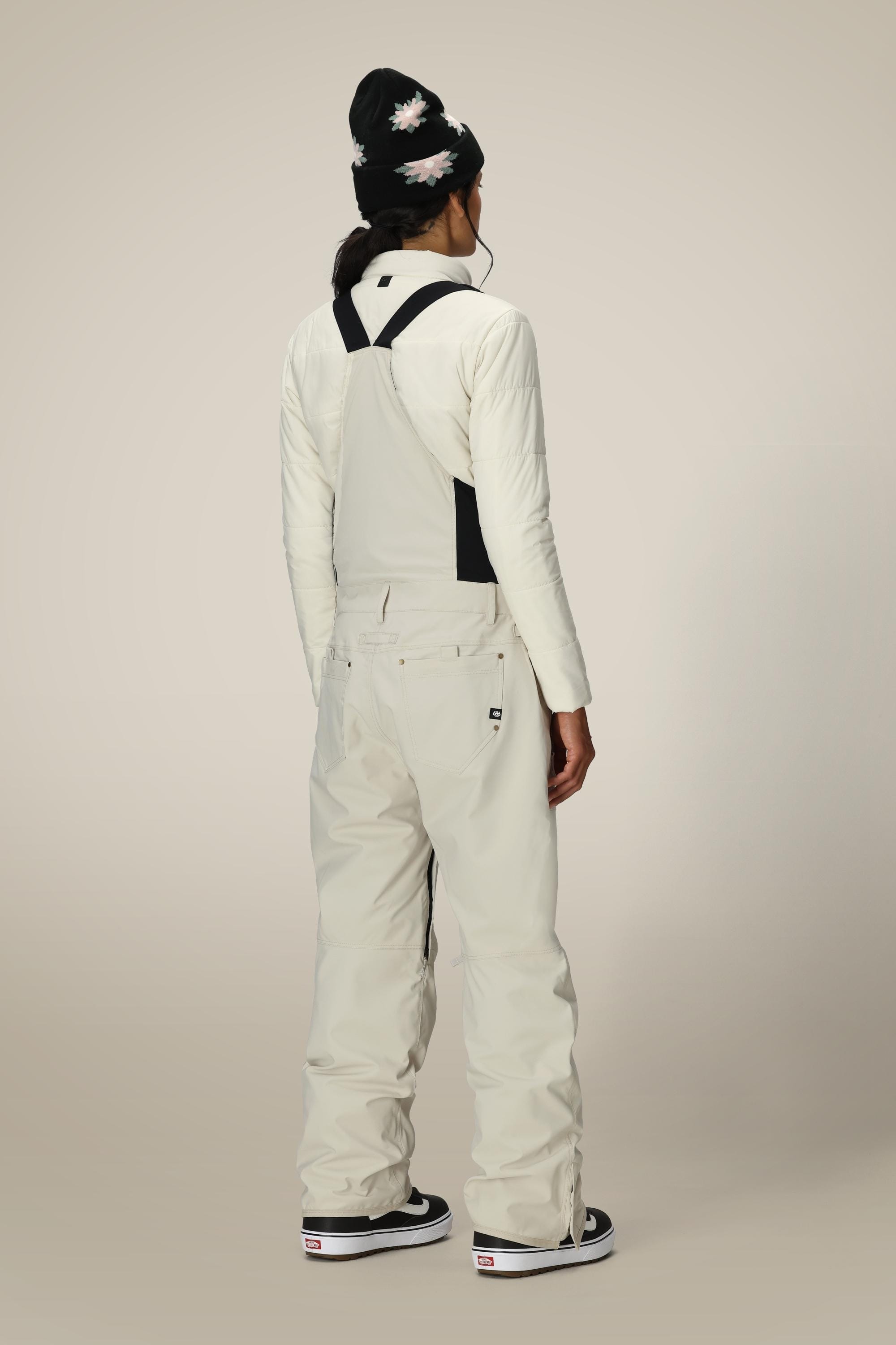 A person stands facing away, wearing the 686 Women’s Black Magic™ Insulated Bib by 686 in off-white with black straps, paired with a long-sleeve shirt, a black beanie featuring a flower pattern, and black sneakers, set against a plain, neutral background.