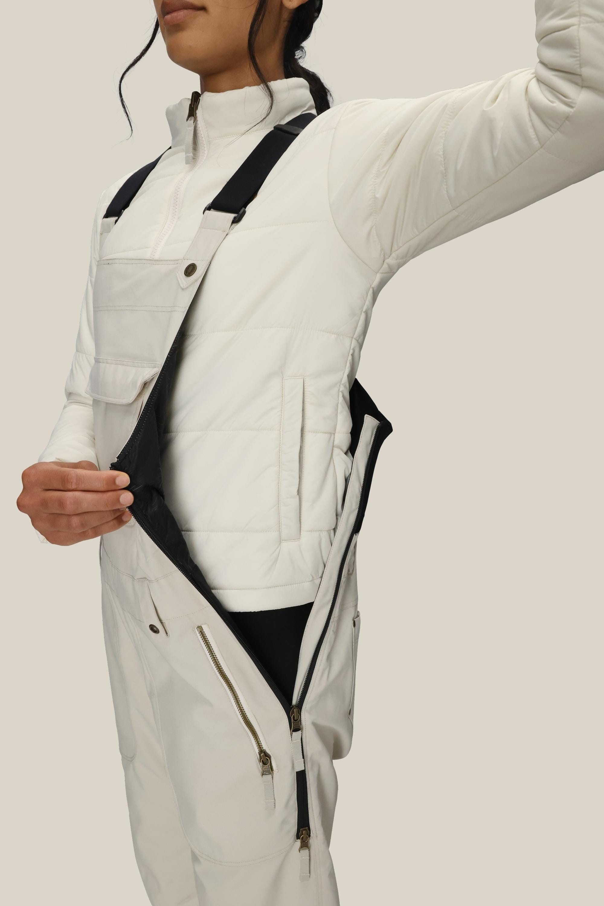 A person wearing the 686 Women’s Black Magic™ Insulated Bib by 686 stands against a plain beige background, unzipping the side zipper to reveal a black inner lining. The bib features black straps and gold zippers.
