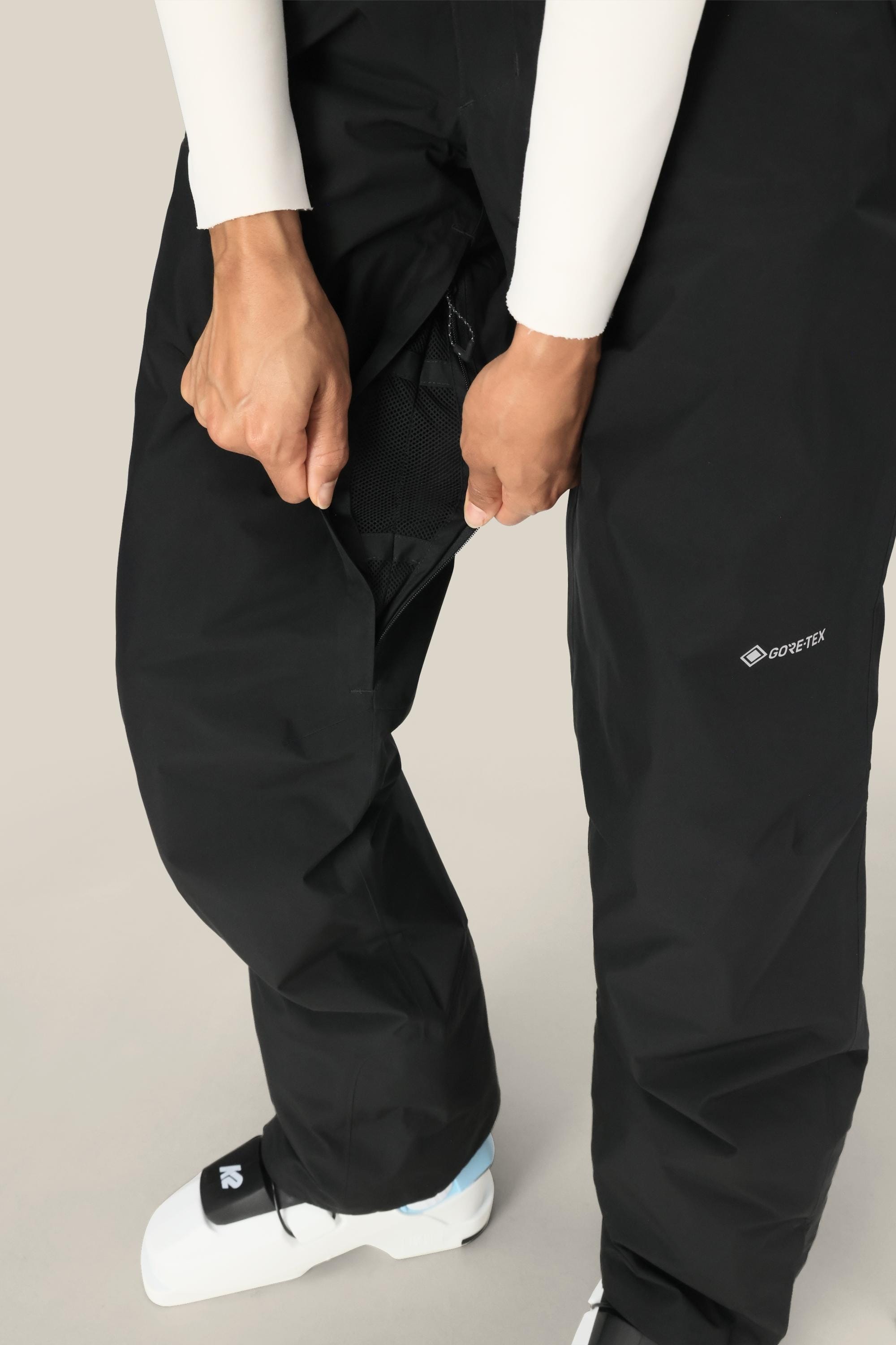 A person wearing 686 Women’s GORE-TEX® Willow™ Insulated Pants and white ski boots unzips a vent on the side of their pants, with only their hands, legs, and lower torso visible.