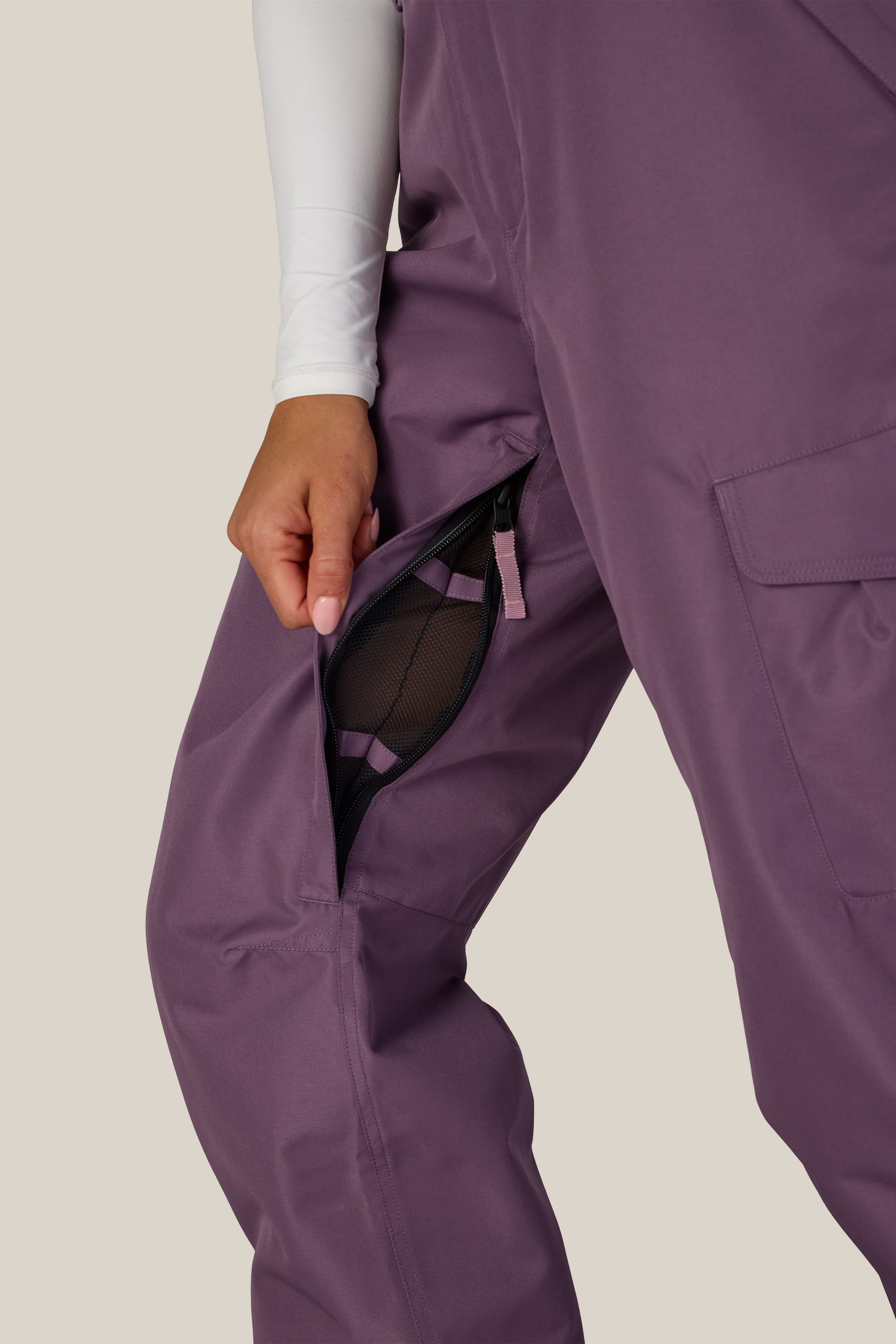 Wearing the 686 Women’s Aura™ Insulated Cargo Pant, a person unzips a mesh-lined side pocket to reveal the interior. Their hand pulls down the zipper while a white long-sleeve shirt from 686 is partially visible.