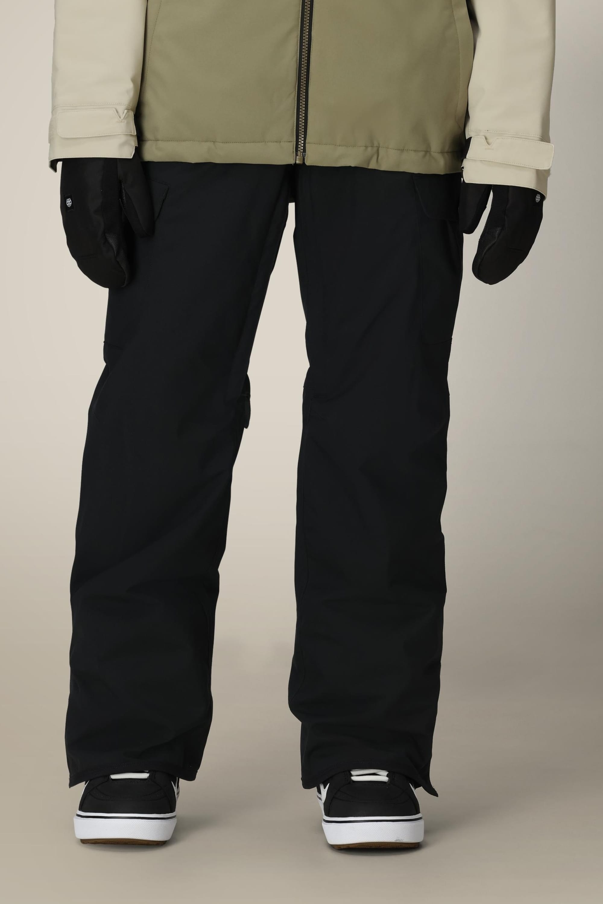 A person wearing 686 Womens Aura Insulated Cargo Pant, black gloves, a beige and white jacket, and black and white shoes stands against a neutral background. The upper body and face are not visible.