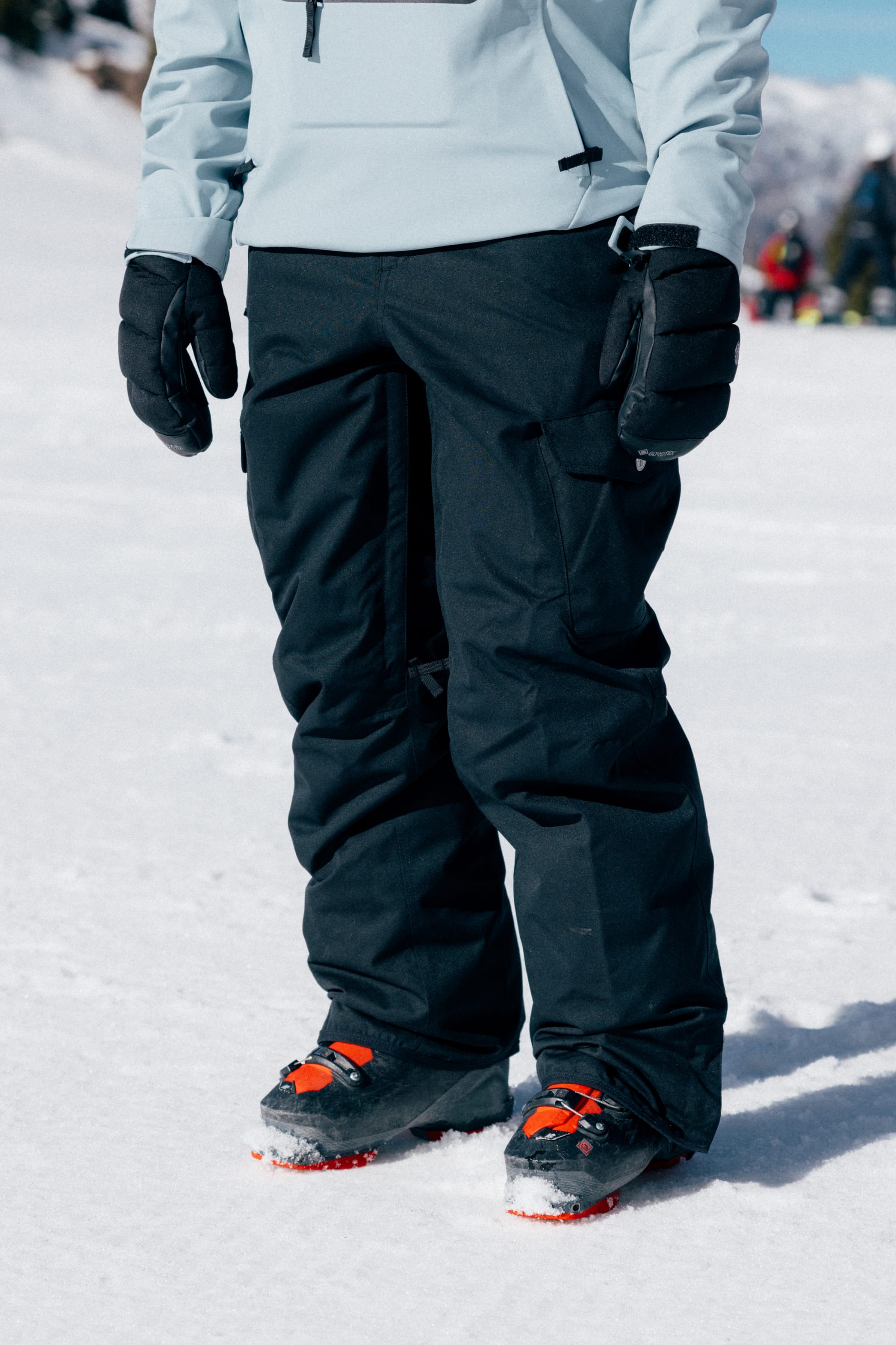A person wearing the 686 Women’s Aura™ Insulated Cargo Pant by 686, along with black gloves and orange-and-black ski boots, stands on snowy ground, with other people and snow-covered terrain visible in the blurred background.