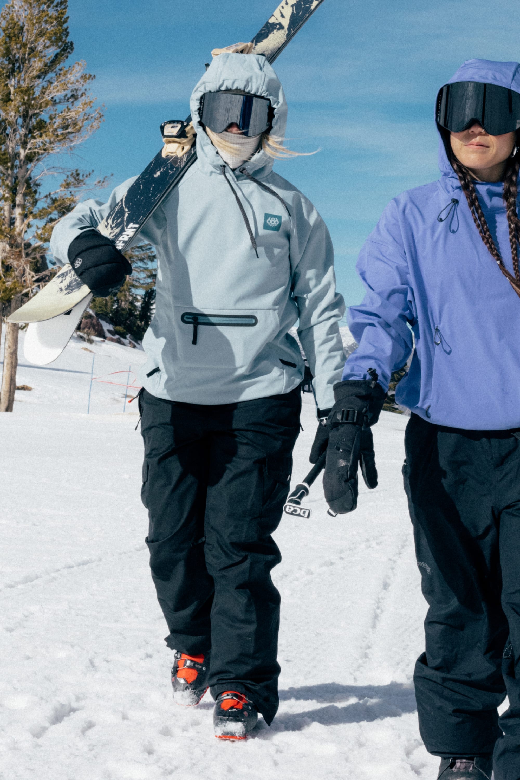 Two people dressed in winter gear trek through the snow—one carries a snowboard on their shoulder, and both wear black goggles and gloves. The person in front is wearing the 686 Women’s Aura™ Insulated Cargo Pant by 686. A tree and blue sky are visible in the background.