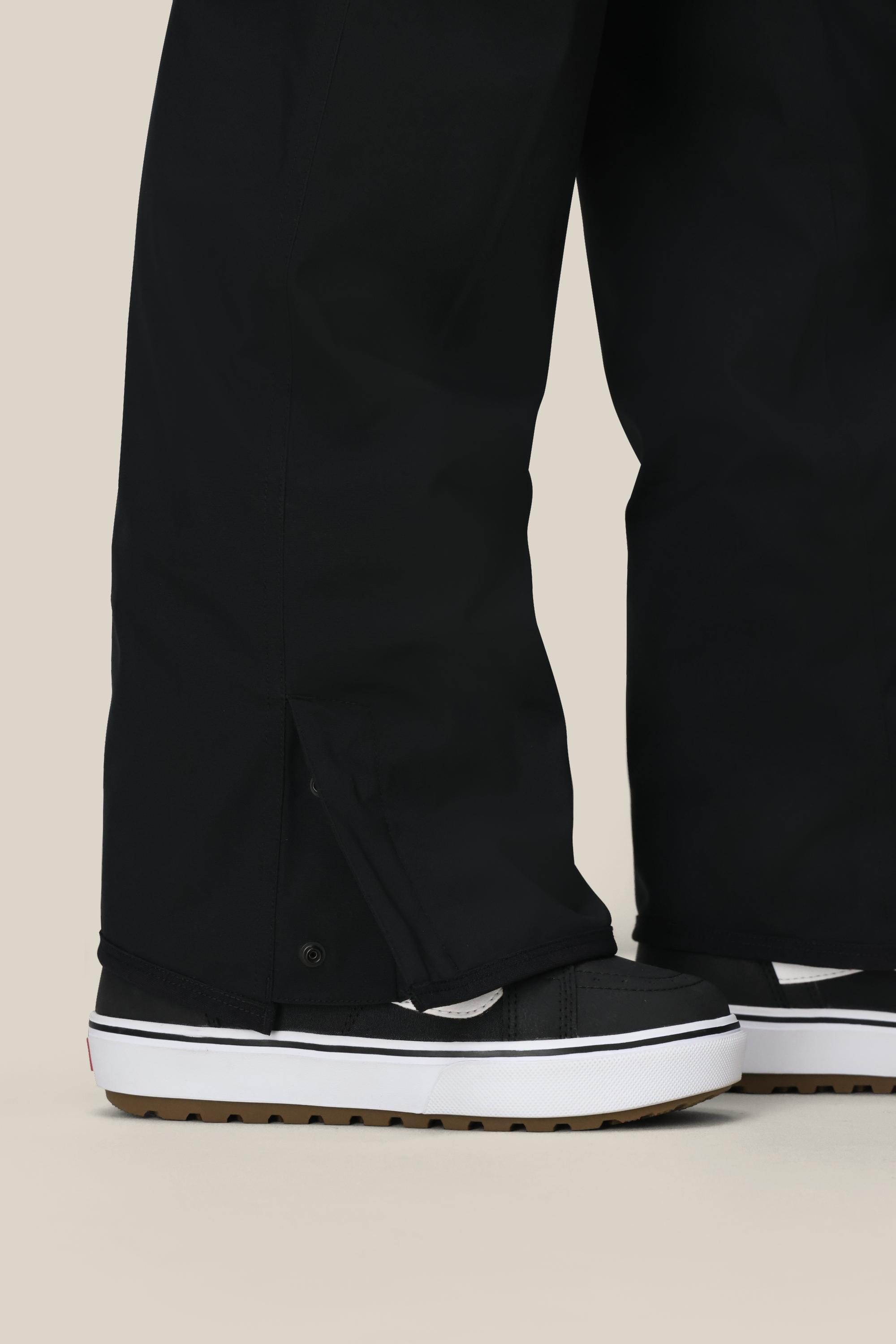 A person stands on a beige background wearing the 686 Womens Aura Insulated Cargo Pant with snap button hems, paired with black and white platform sneakers.