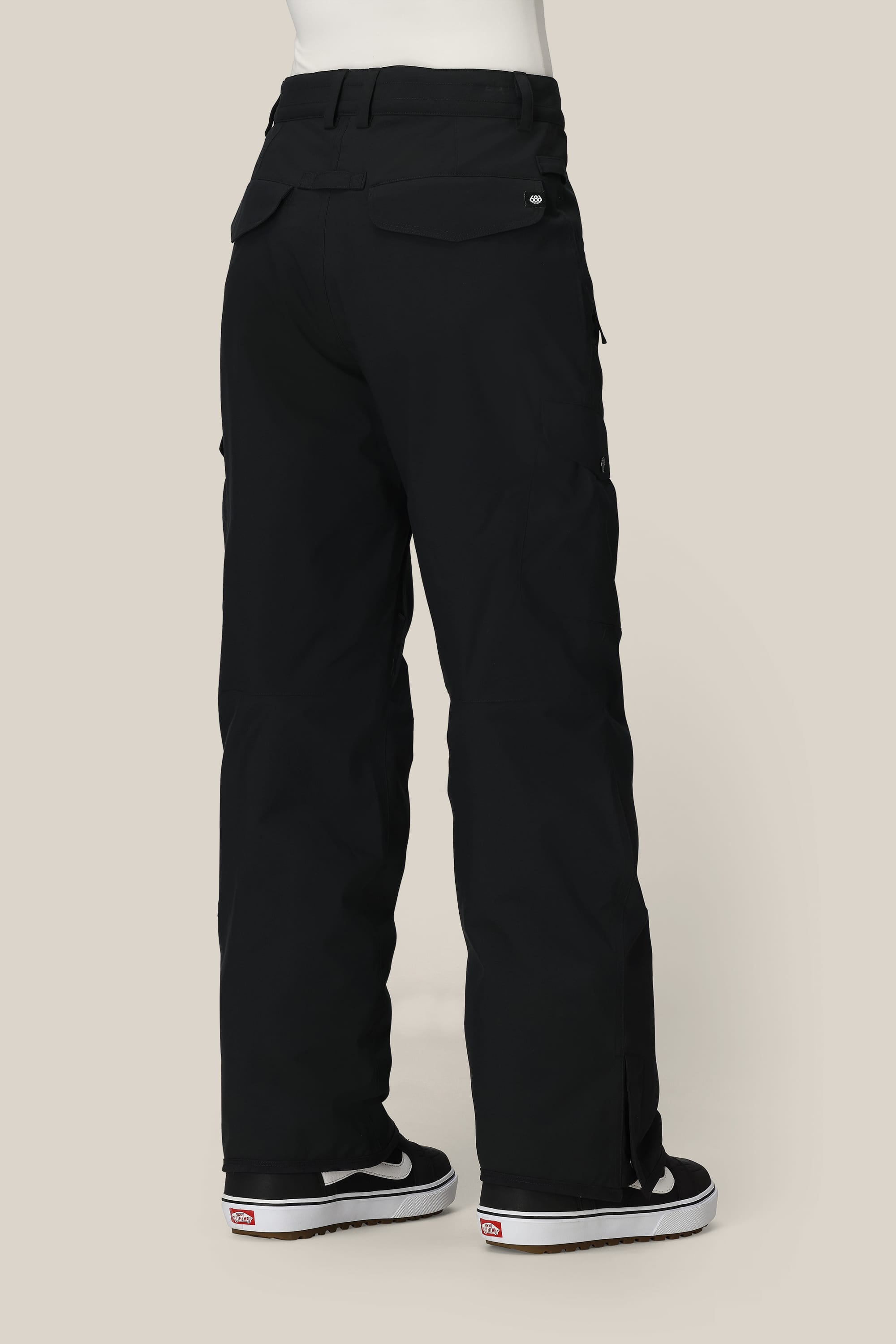 A person wearing 686 Womens Aura Insulated Cargo Pant and black-and-white Vans shoes stands facing away from the camera against a plain beige background.