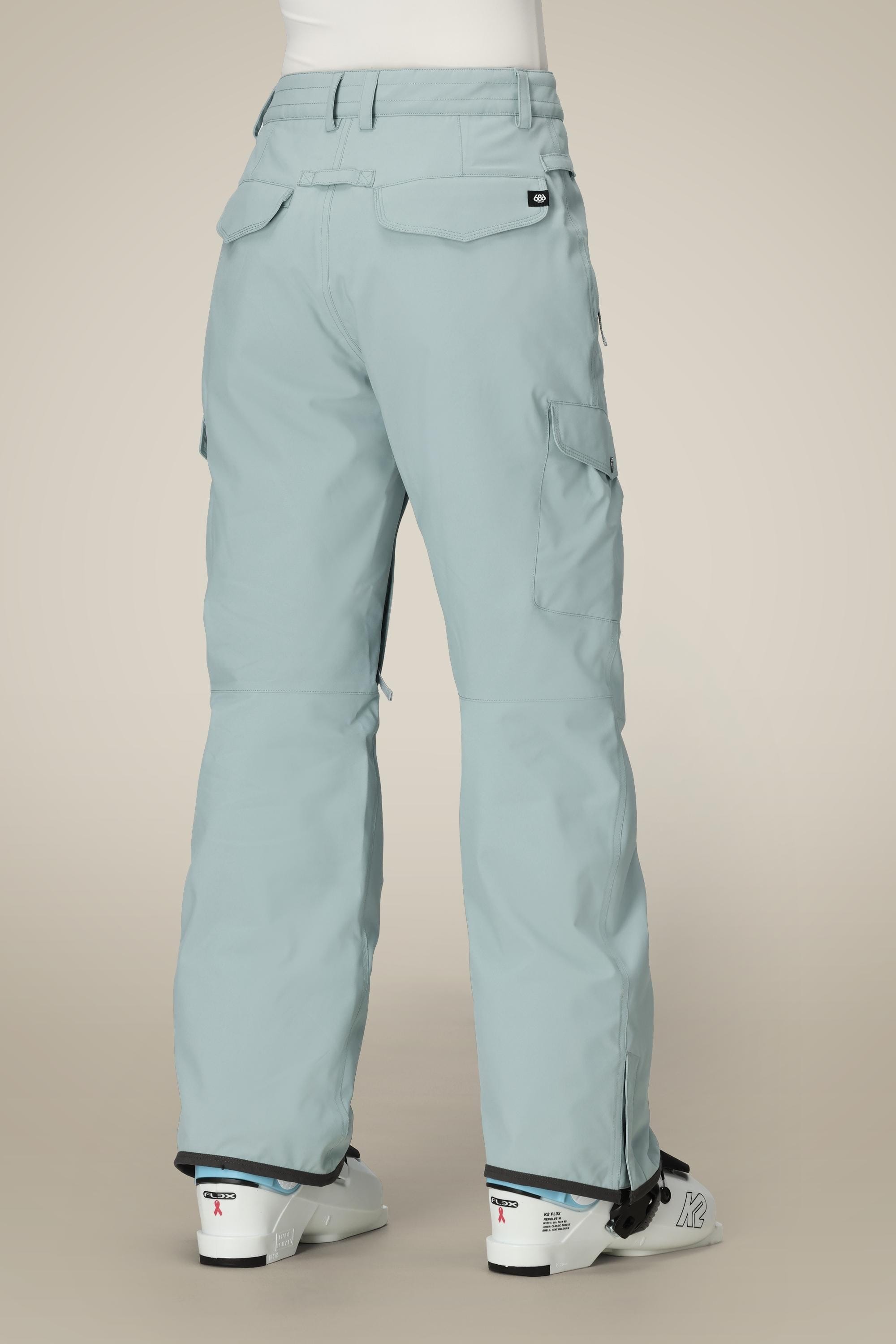 Person wearing 686 Womens Aura Insulated Cargo Pant in light blue with white ski boots, shown from the back. These waterproof polyester pants by 686 feature flap pockets and a relaxed fit ideal for winter sports.