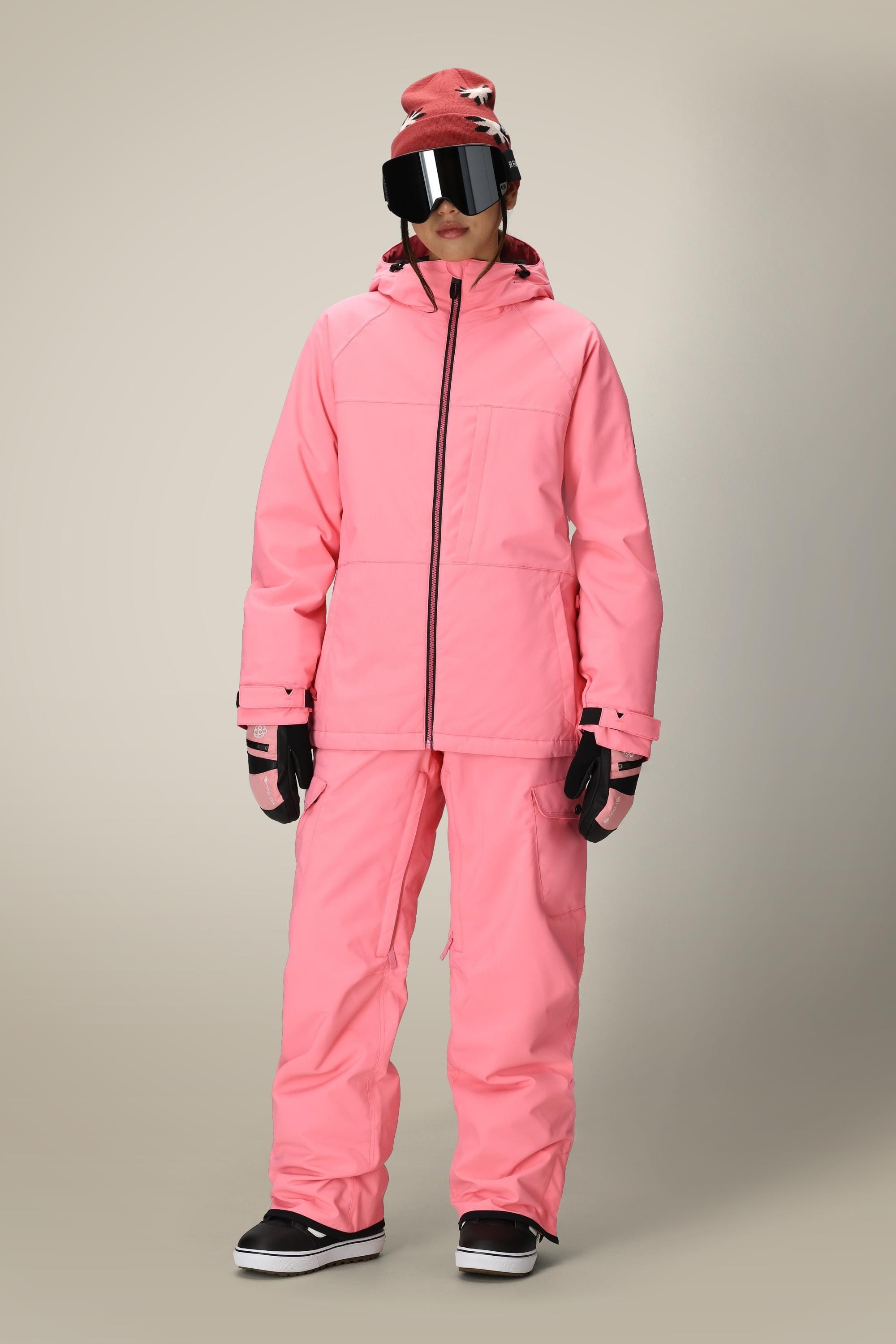 A person wearing a bright pink ski jacket and 686 Womens Aura Insulated Cargo Pant, black gloves, black snow boots, large reflective ski goggles, and a pink beanie stands facing forward against a plain background.