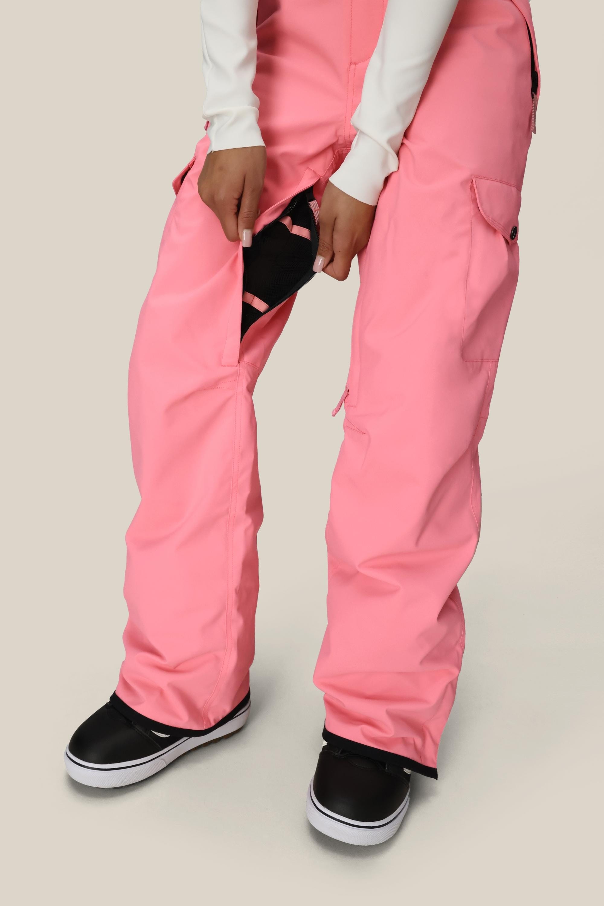 A person wearing 686 Womens Aura Insulated Cargo Pant in pink and black sneakers stands on a light background, unzipping the side vent to reveal the black inner lining, with hands gripping the zipper and fabric.