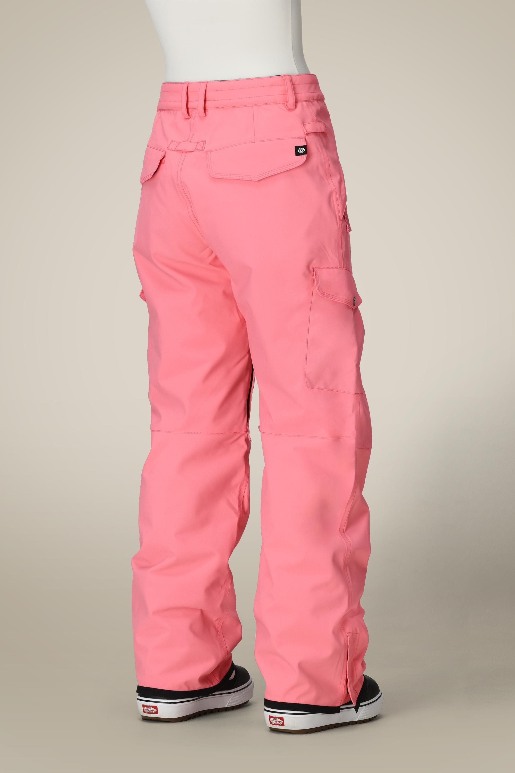 A mannequin showcases the 686 Womens Aura Insulated Cargo Pant in bright pink, featuring multiple pockets, paired with black-and-white sneakers and displayed from the back against a plain beige background.