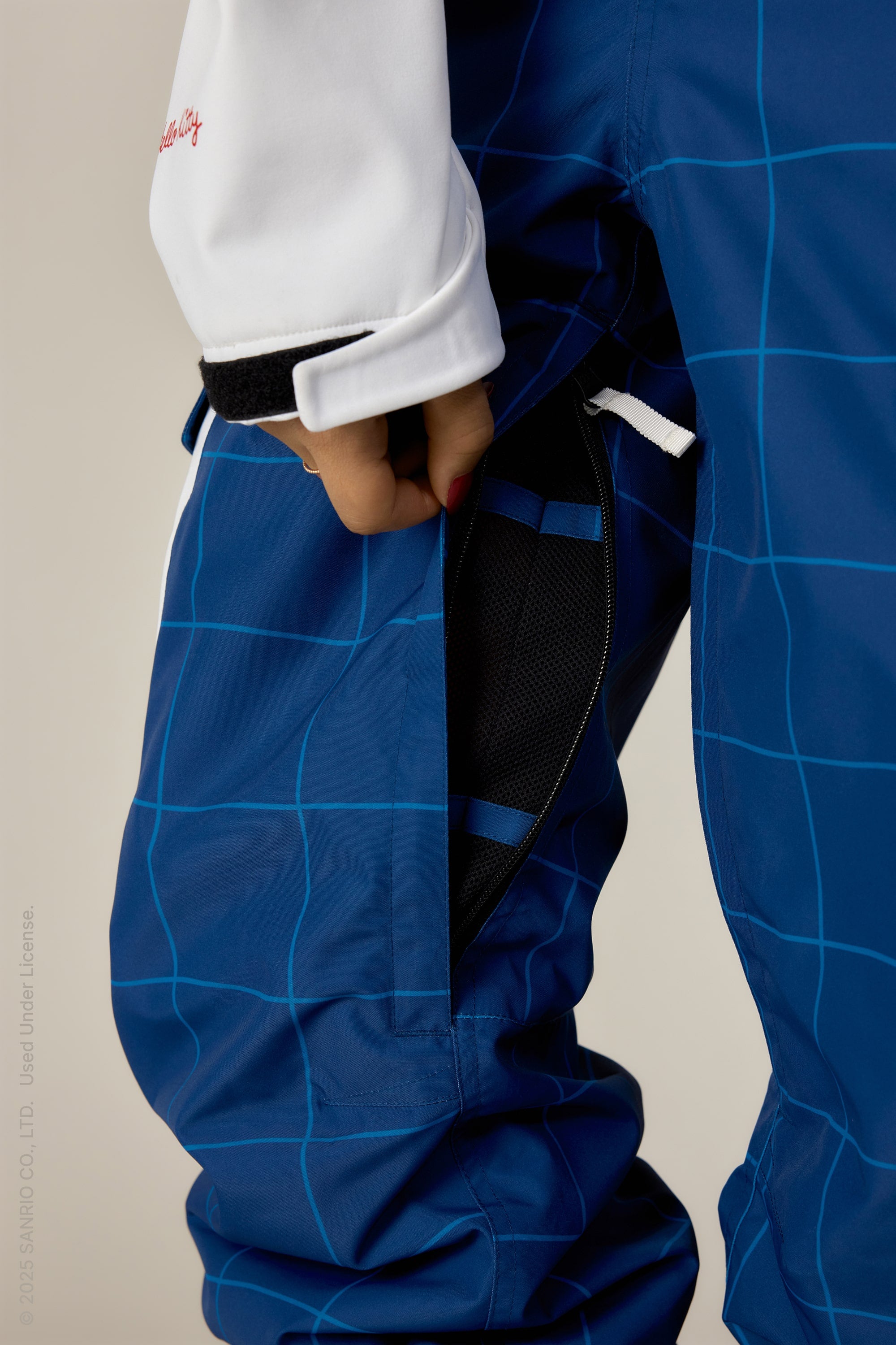 A person wearing 686 Womens Aura Insulated Cargo Pant with a light blue grid pattern and a white sleeve opens a zippered side vent, showing the black mesh lining underneath.