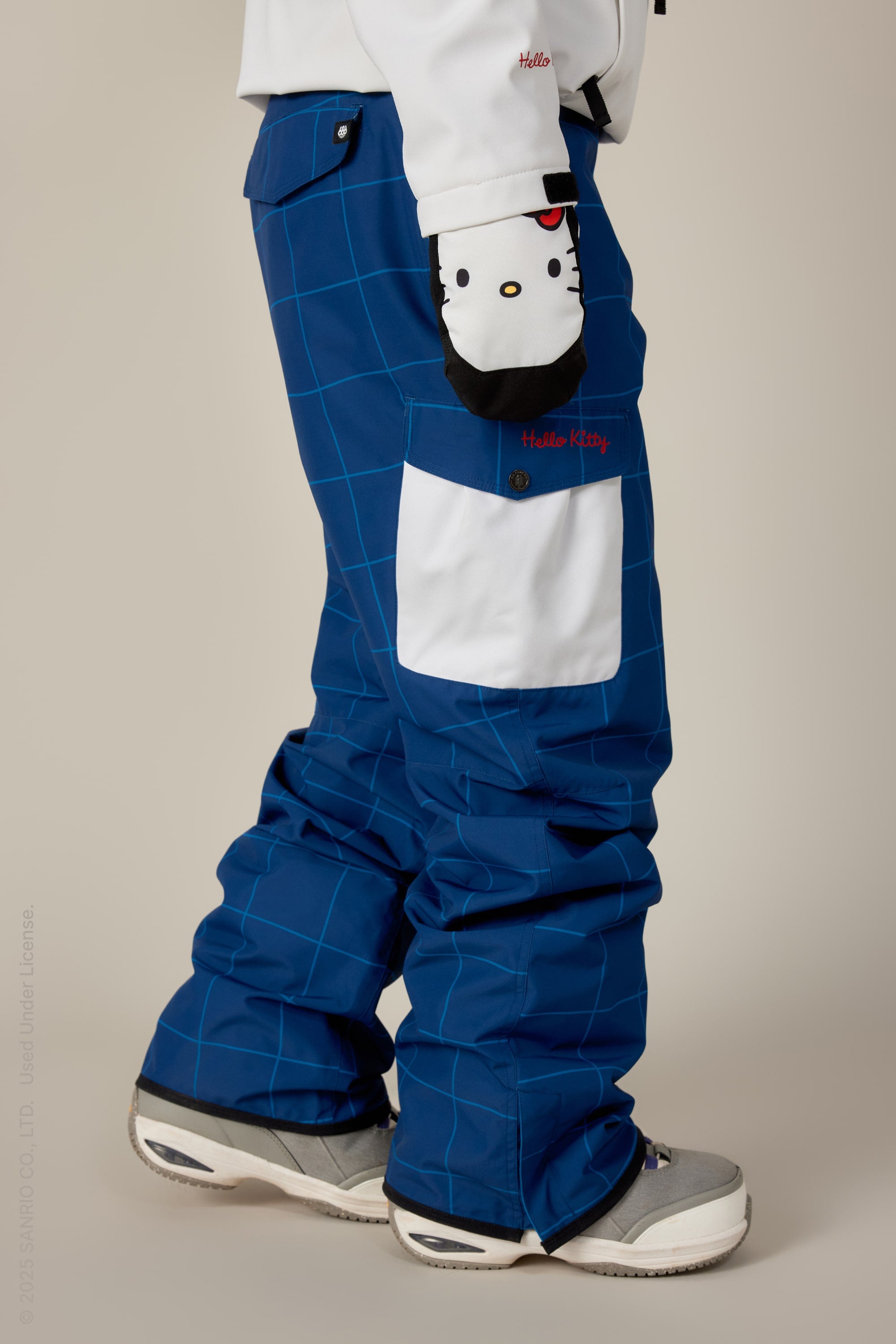 A person wears 686 Women’s Aura Insulated Cargo Pants in blue check, a white jacket, gray and white sneakers, and a Hello Kitty glove. The pants have a large white Hello Kitty-labeled pocket.