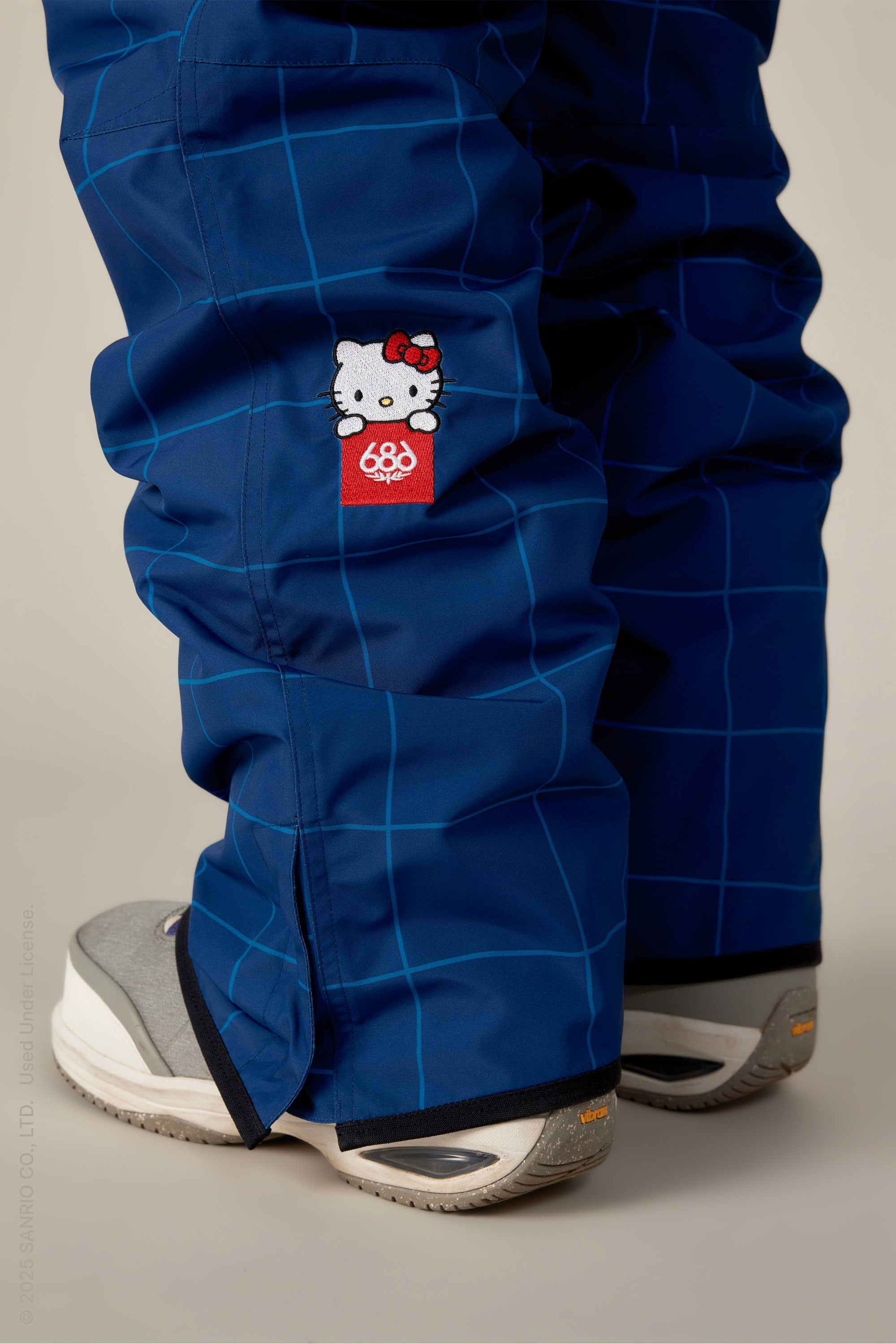 The 686 Womens Aura Insulated Cargo Pant, shown in blue with a light blue grid pattern and Hello Kitty patch above a red 686 logo, is styled with gray and white athletic shoes. Only the lower legs and feet are visible against a neutral background.
