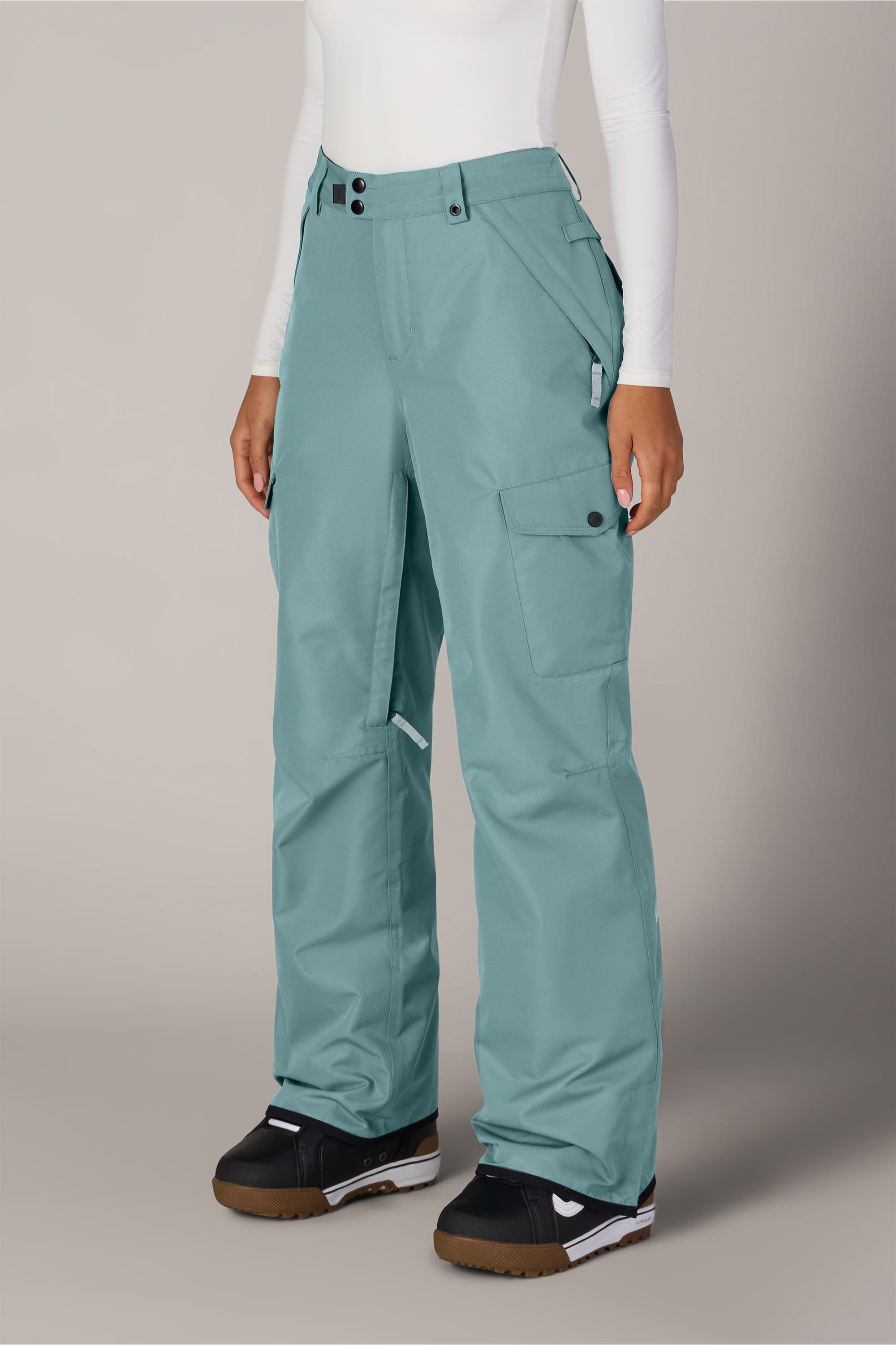 A person wears the 686 Women’s Aura™ Insulated Cargo Pant in light blue, paired with a white long-sleeve top and black snow boots, standing against a plain, light background. Only the lower body is visible.