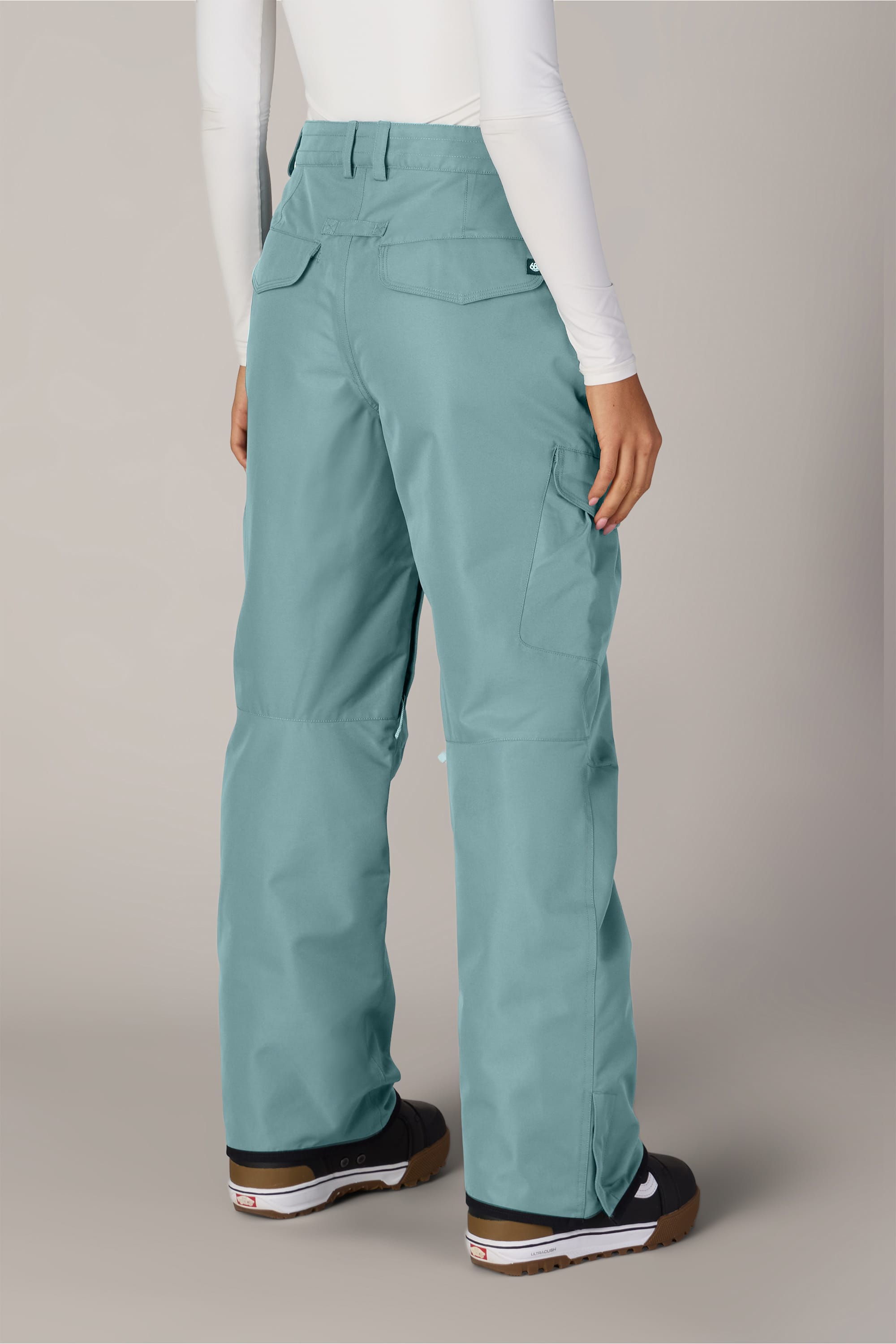 Wearing the 686 Women’s Aura™ Insulated Cargo Pant in teal, paired with a white long-sleeve top and black boots, a person stands facing away to showcase the back view of the outfit.