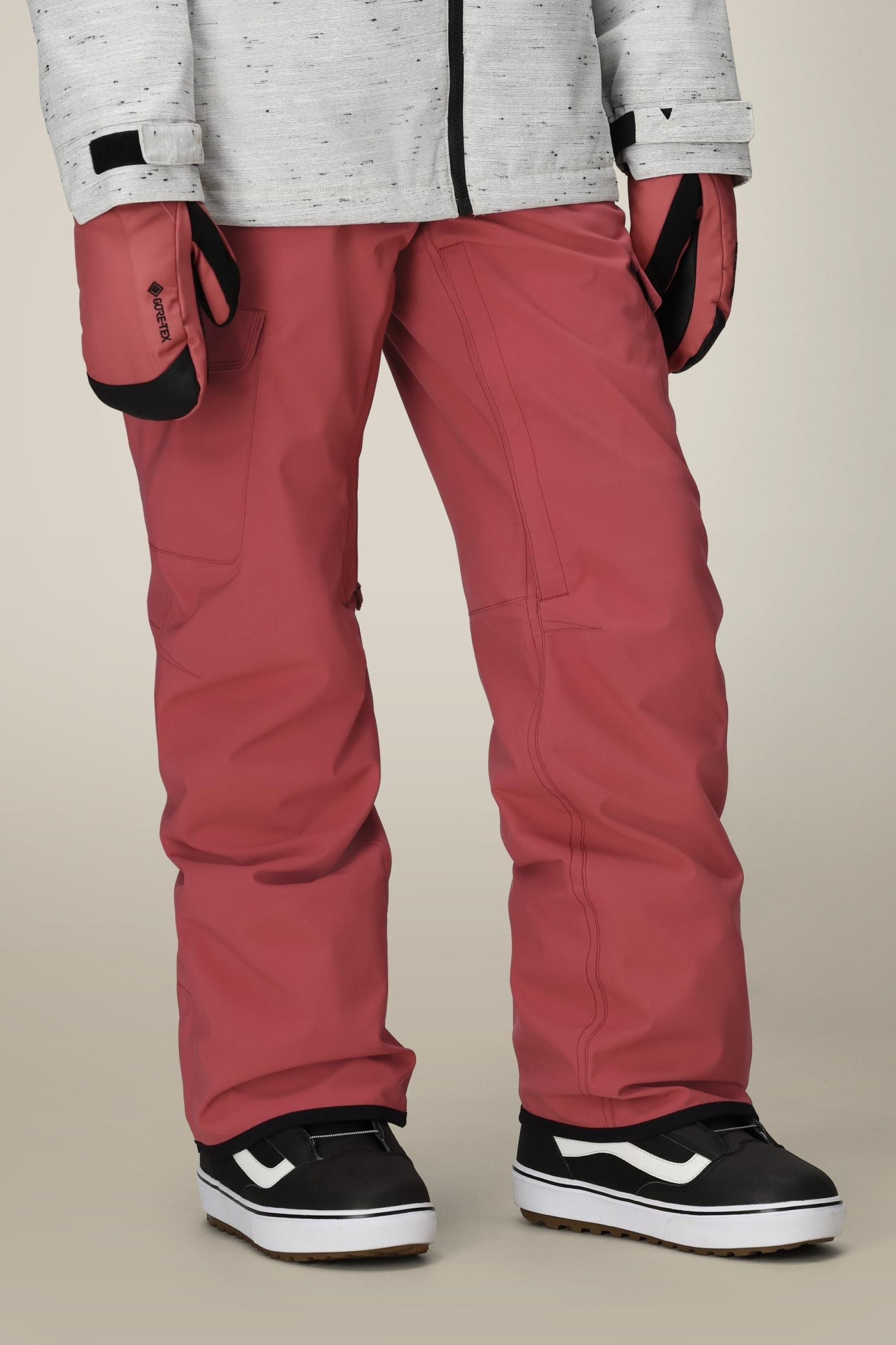 Wearing the 686 Womens Aura Insulated Cargo Pant in red, paired with a light gray jacket, red gloves, and black-and-white sneakers, a person stands against a neutral background. Only their lower half is visible.