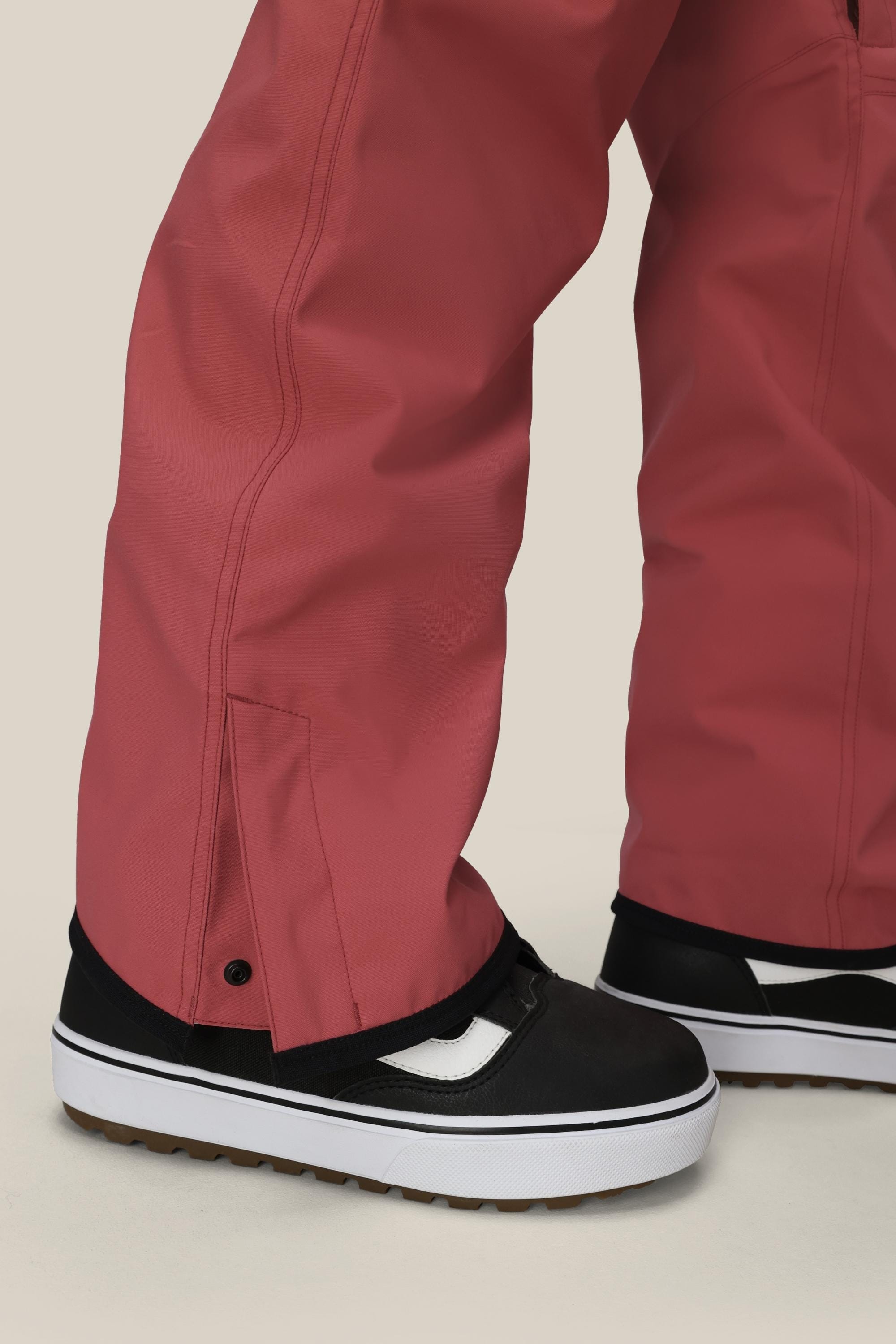 A close-up of a person wearing 686 Womens Aura Insulated Cargo Pant by 686, styled loose in rose and covering most of their black-and-white skate shoes, set against a light beige background.