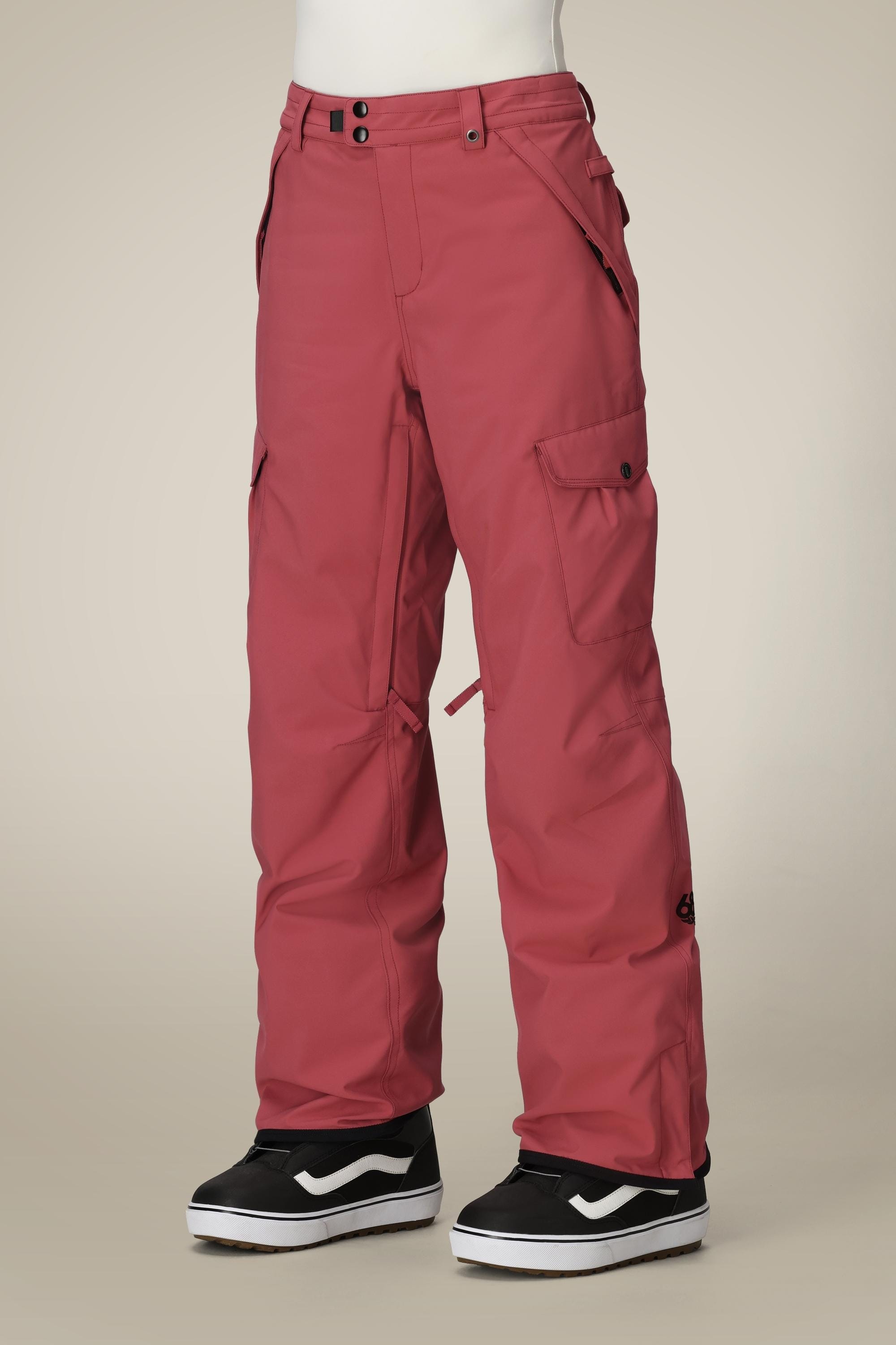 A person wears 686 Womens Aura Insulated Cargo Pant in red and black-and-white Vans sneakers, standing against a plain beige background. The 686 cargo pants feature multiple pockets and button closures.