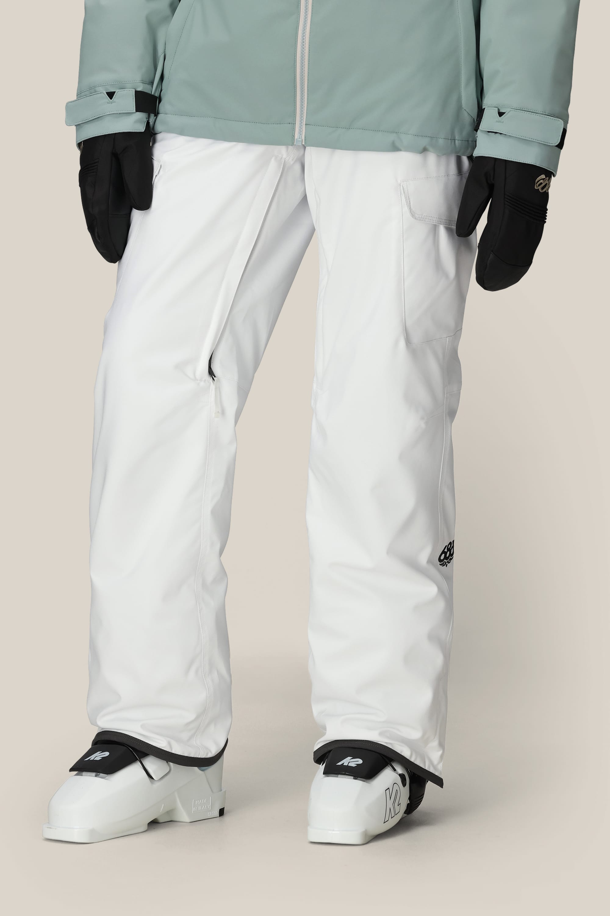 A person is wearing 686 Womens Aura Insulated Cargo Pants in white, black gloves, white ski or snowboard boots, and a light green jacket. The background is plain and neutral.