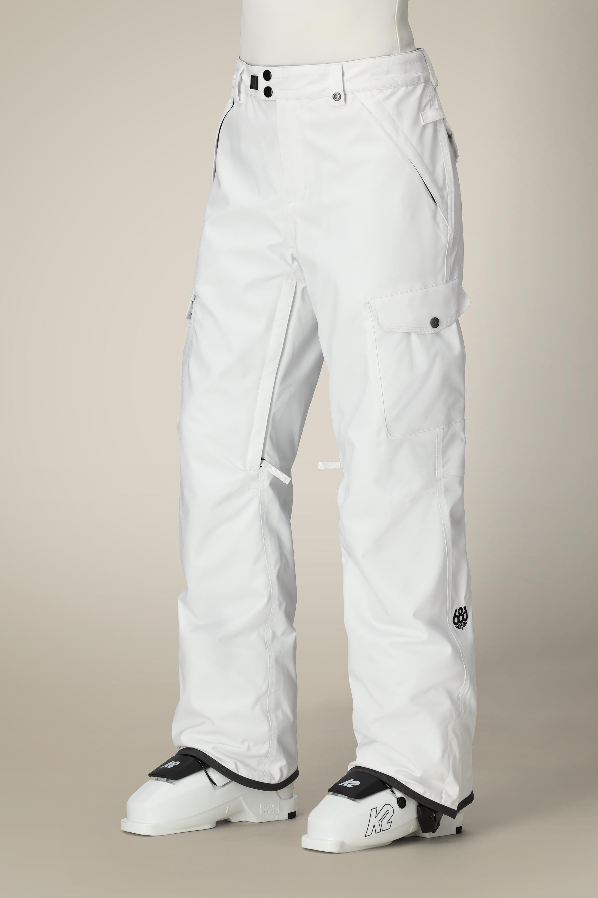 A person wears 686 Womens Aura Insulated Cargo Pant—loose-fitting, white, with black buttons and pockets—paired with white ski or snowboard boots featuring black accents, standing against a plain background.