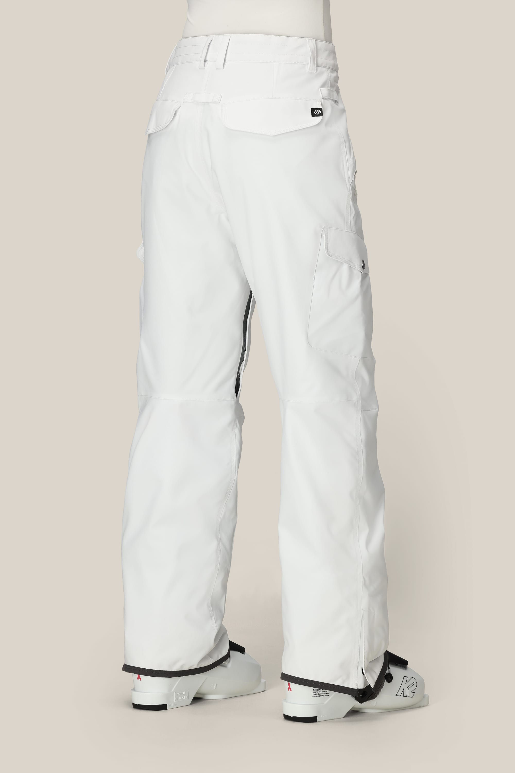 A person is shown from the back wearing white 686 Womens Aura Insulated Cargo Pant and white ski boots, against a plain light beige background. The waterproof polyester pants by 686 feature back and side cargo pockets with flap closures.