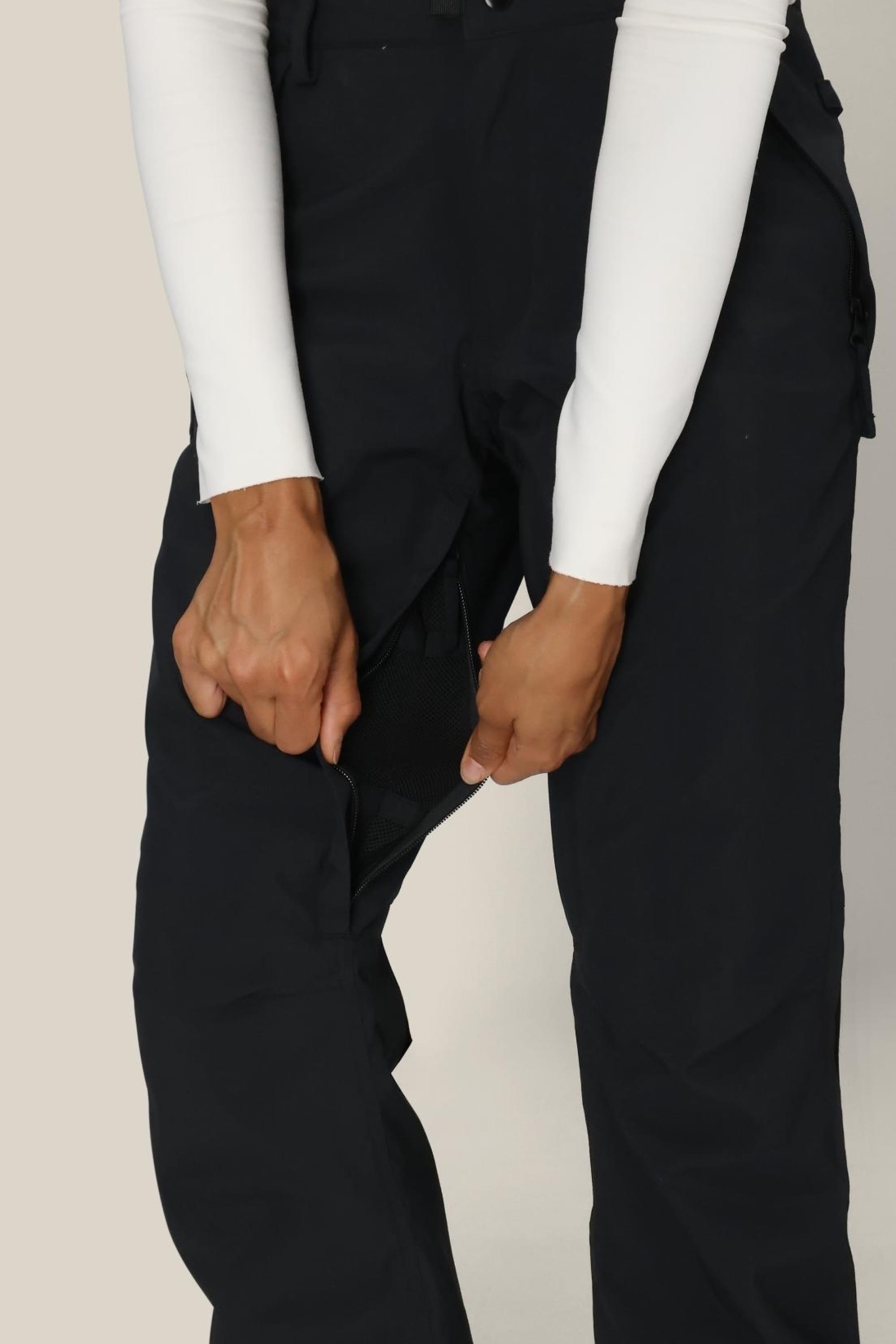 A person wearing the 686 Women’s Standard Shell Pant in black and a white long-sleeve top uses both hands to unzip a concealed zipper on the inner thigh of the pants, revealing a mesh lining beneath.