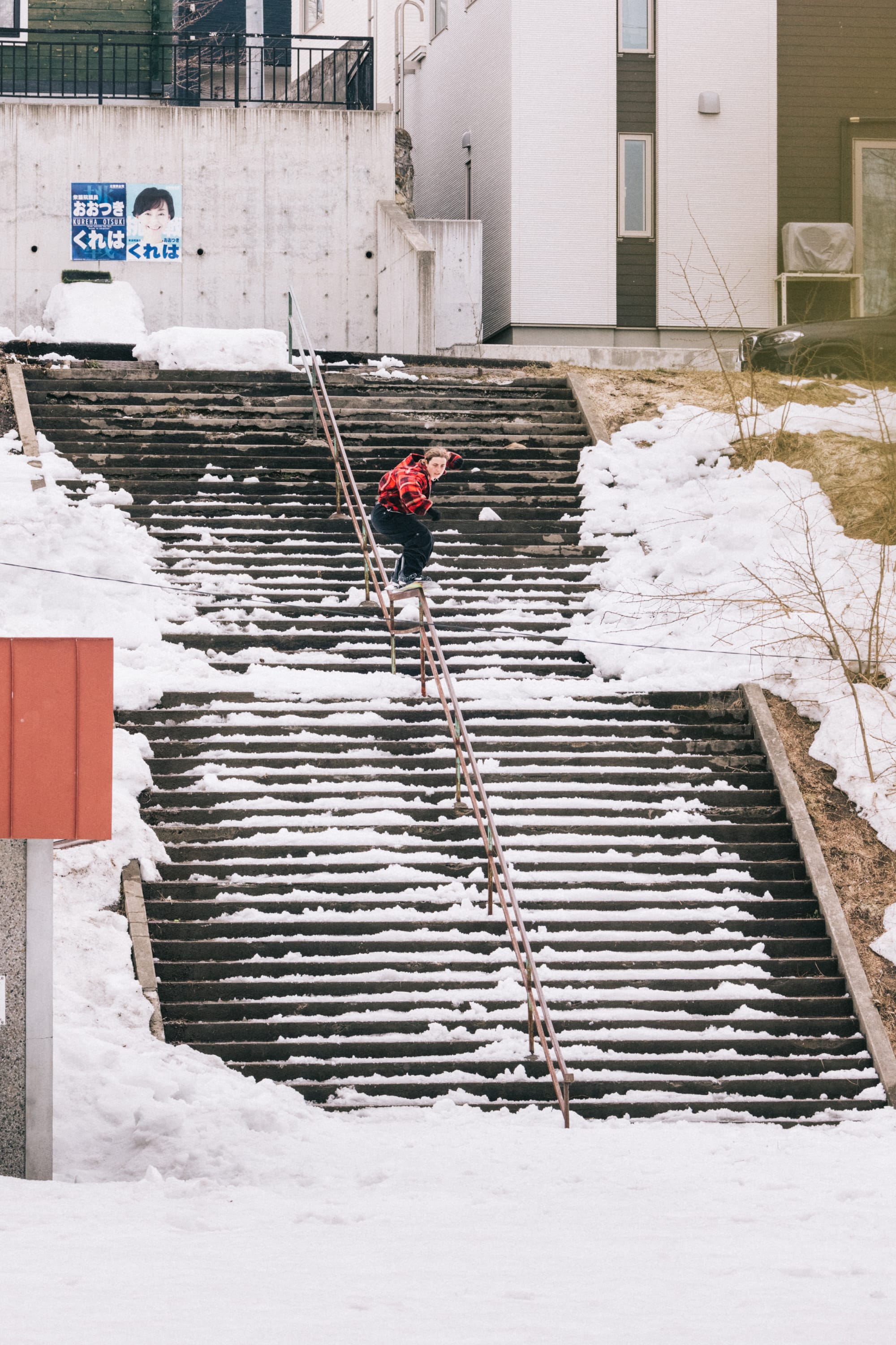 A person wearing the 686 Women’s Outline™ Shell Pant by 686 snowboards down a handrail between snowy steps outside, dressed in a red jacket and helmet, with snow-covered ground and buildings all around.