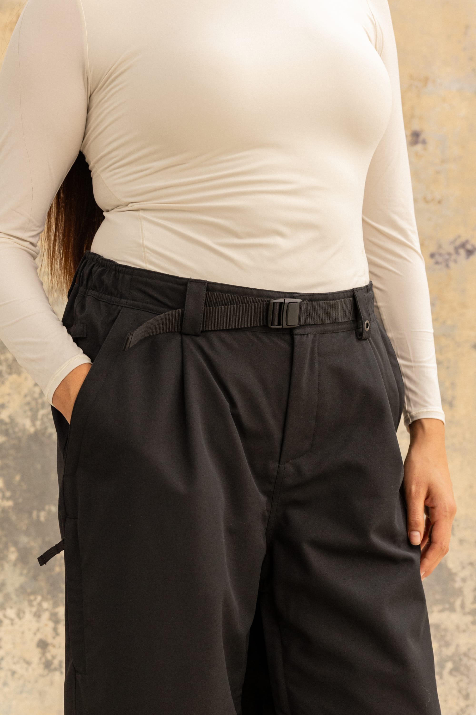 A person wearing a fitted long-sleeve beige top and the 686 Women’s Outline™ Shell Pant by 686—a loose black pant with a belt—is standing with one hand in their pocket against a textured beige background.