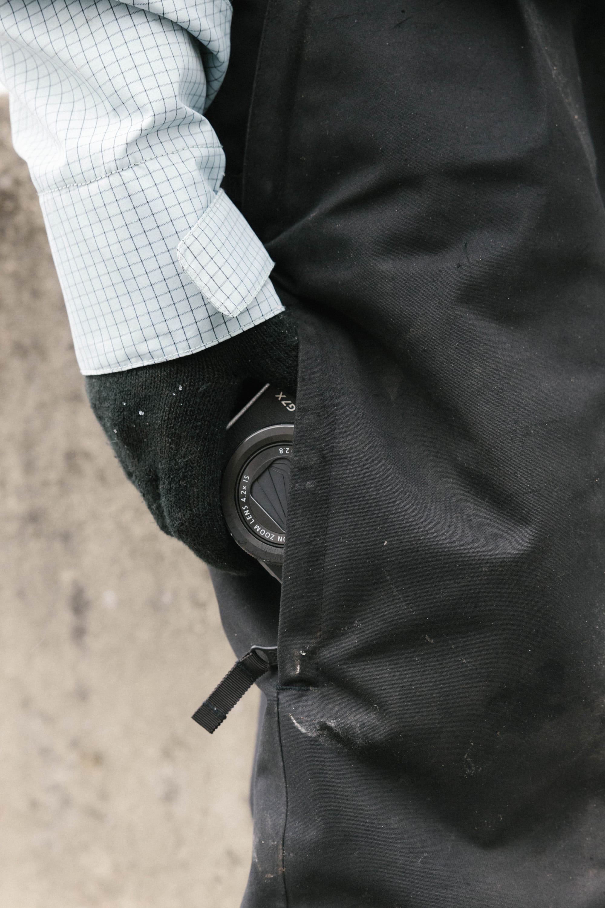 A person wearing a light blue checkered shirt and black glove has a camera lens partially tucked into the pocket of 686 Women’s Outline™ Shell Pant by 686.