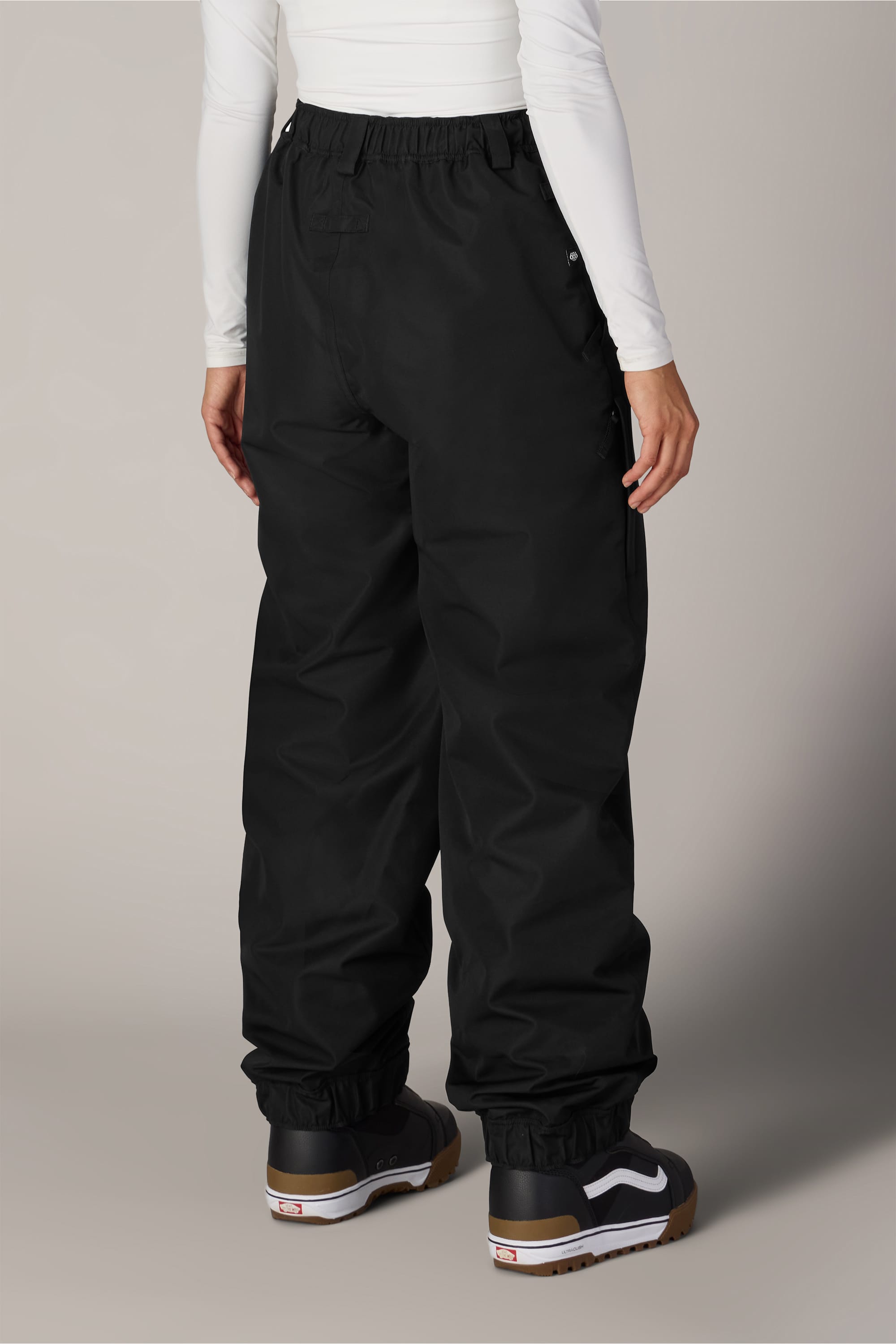 A person wearing the 686 Women’s Outline™ Shell Pant in black, paired with a fitted white long-sleeve top and black Vans sneakers, is standing against a plain background, shown from the back.