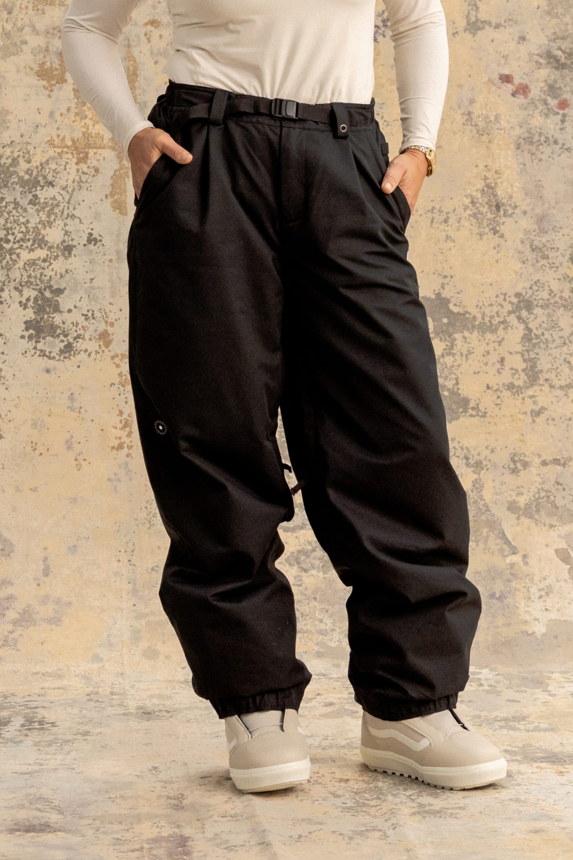 A person wears the 686 Women's Outline™ Shell Pant by 686 in black, paired with a long-sleeve white top, white chunky shoes, and a gold watch, standing with hands in pockets against a textured beige and gray backdrop.