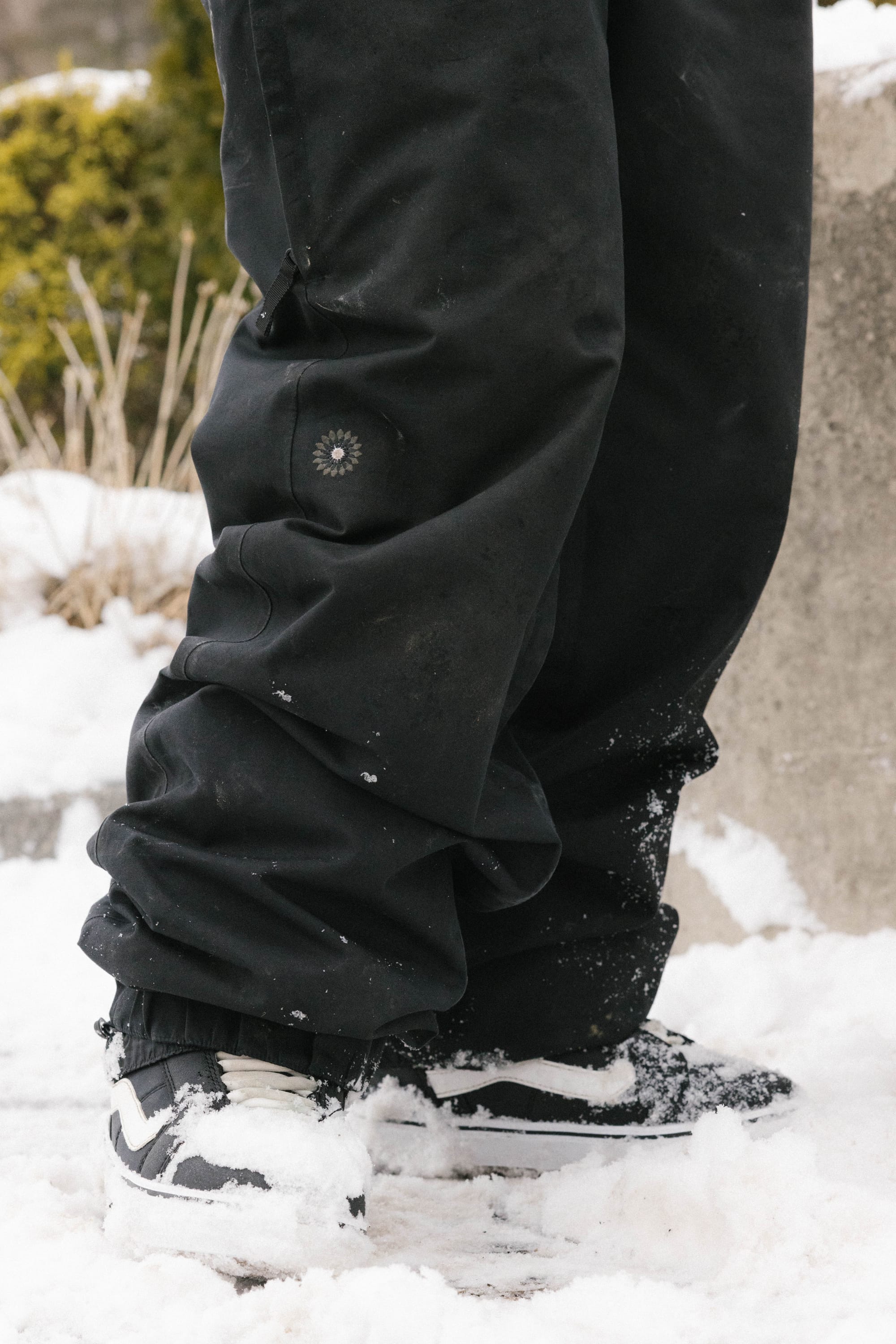 A person wearing 686 Women's Outline™ Shell Pant by 686 and black-and-white sneakers stands on snow-covered ground, with green plants blurred in the background.