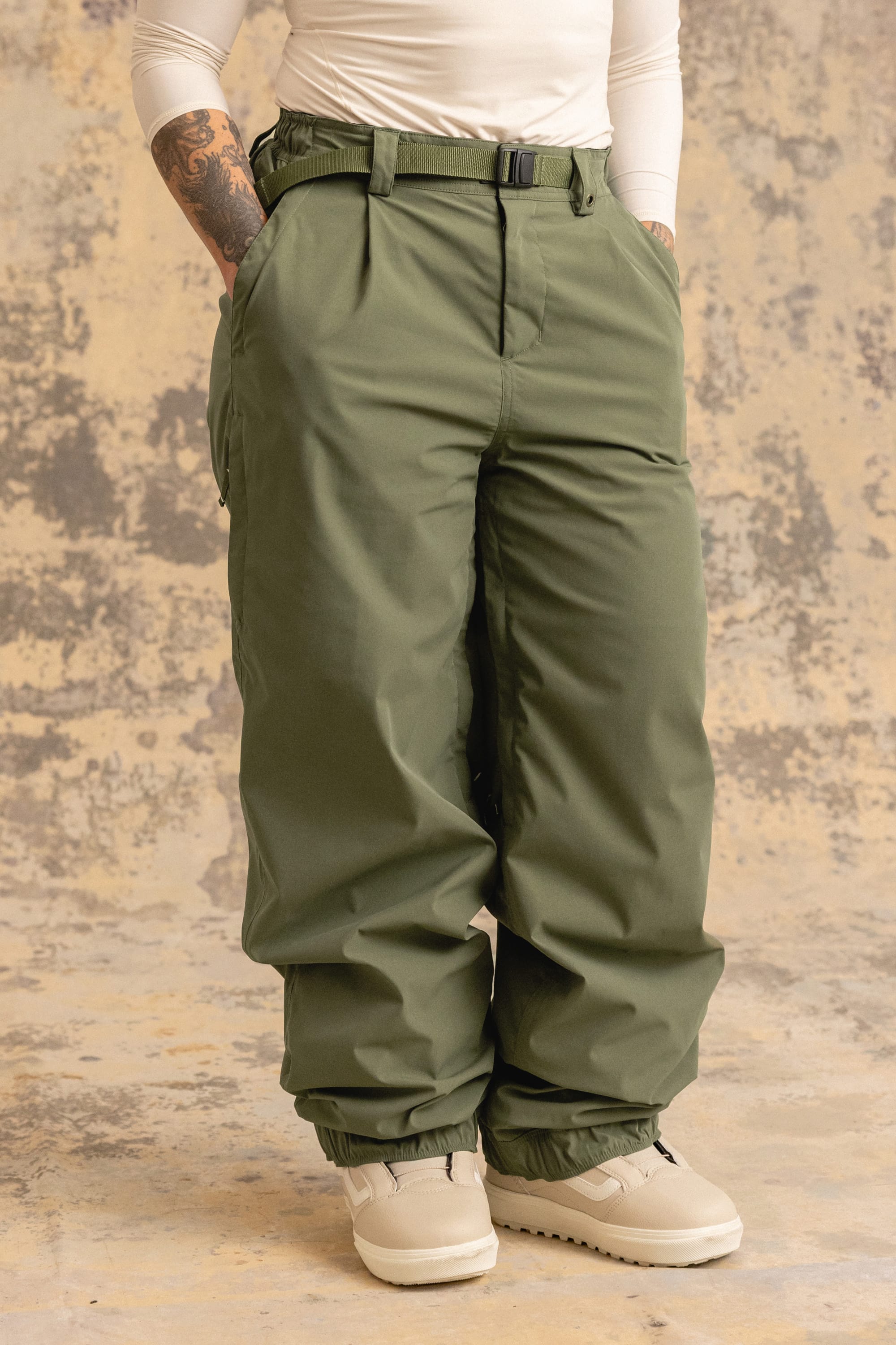A person stands with hands in pockets against a mottled beige background, wearing the 686 Women’s Outline™ Shell Pant from 686 in olive green. She pairs the pants with white shoes and a white long-sleeve shirt, and tattoos are visible on one forearm.