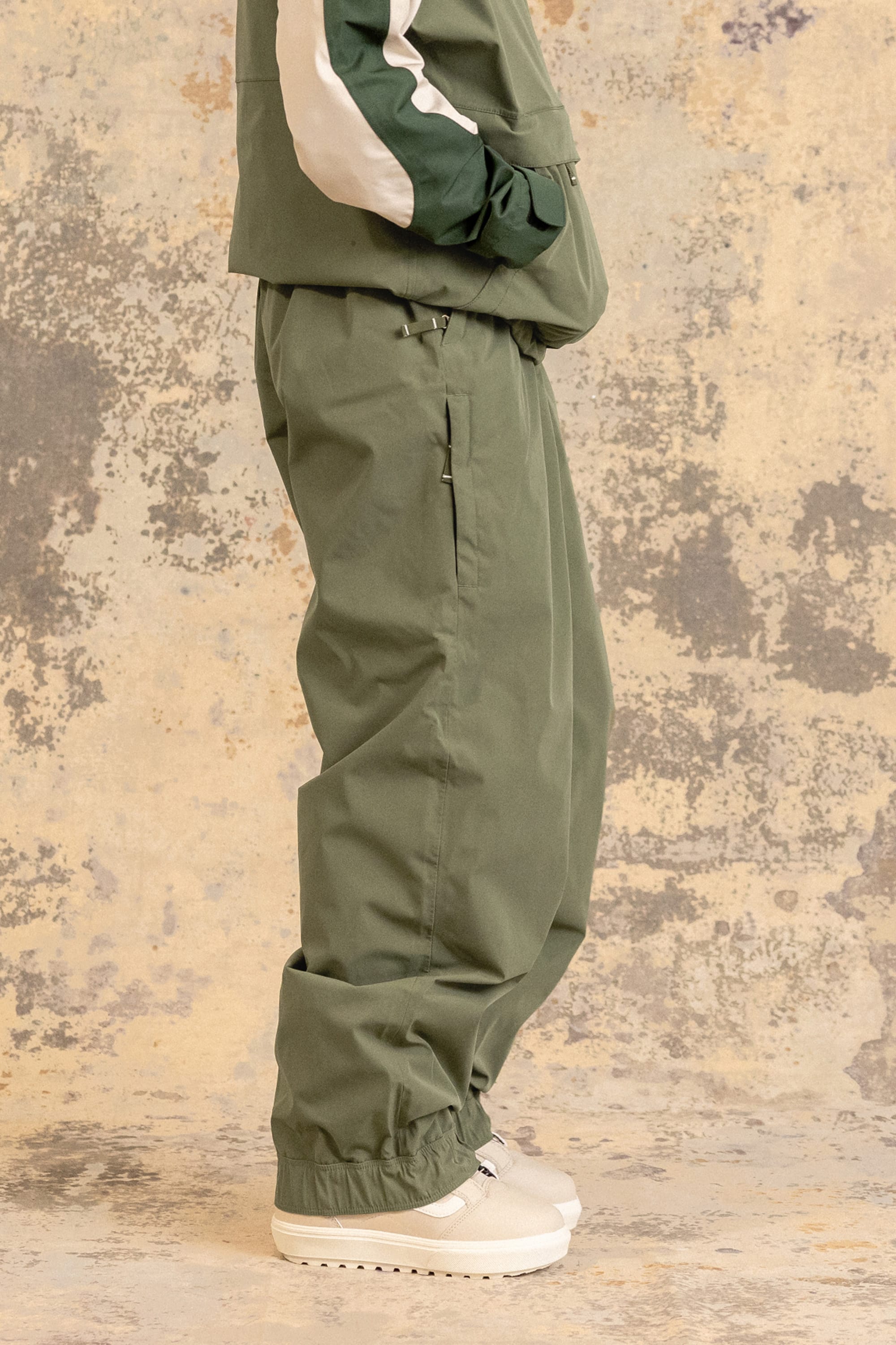 A person stands sideways wearing the 686 Women’s Outline™ Shell Pant in green, paired with a matching green jacket featuring a white stripe and white sneakers. The background is textured with beige and gray tones.