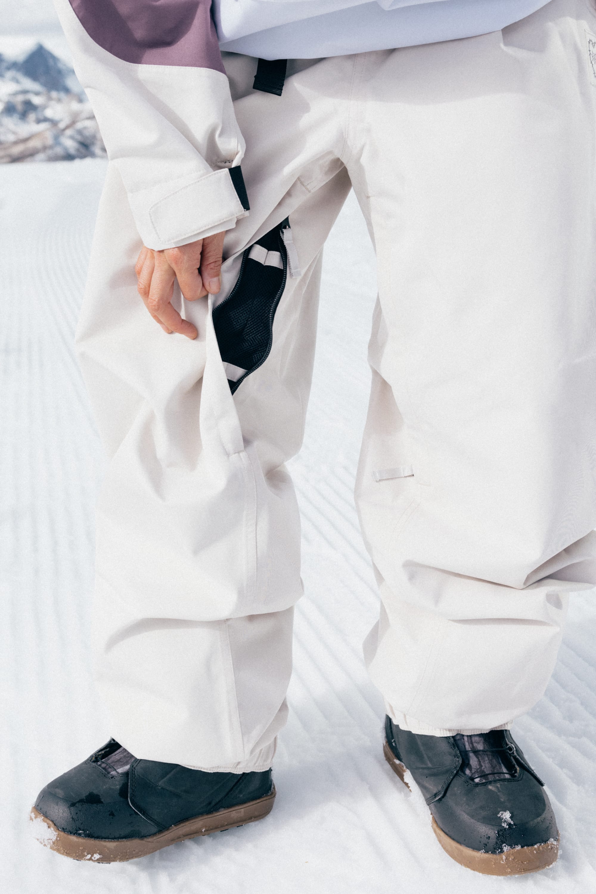 A person is standing on snow in 686 Women’s Outline™ Shell Pant by 686, unzipping the side pocket to reveal black fabric inside, with snowy mountains softly blurred in the background.