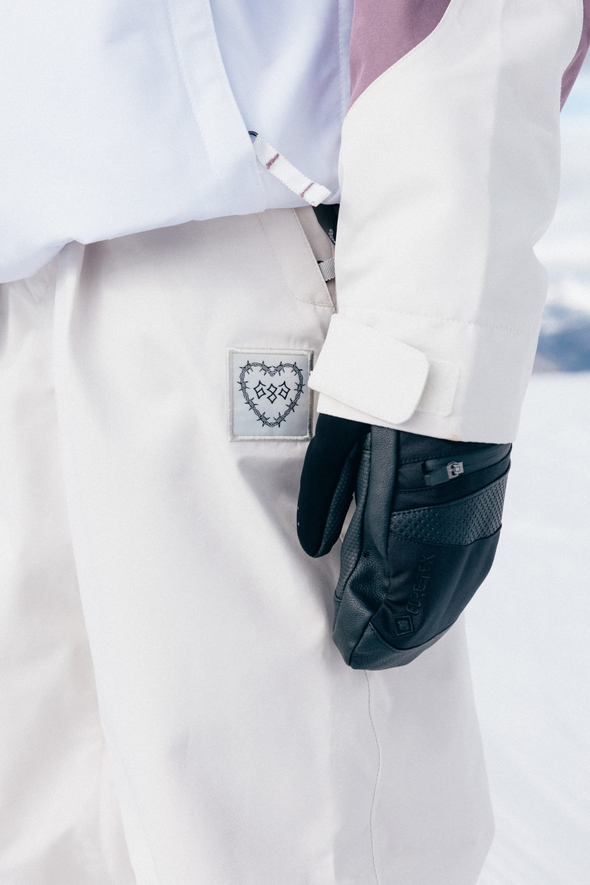 A person is wearing the 686 Women’s Outline™ Shell Pant along with a jacket featuring a visible patch with a heart and barbed wire design. They are also wearing black gloves and standing outdoors on a snowy surface.