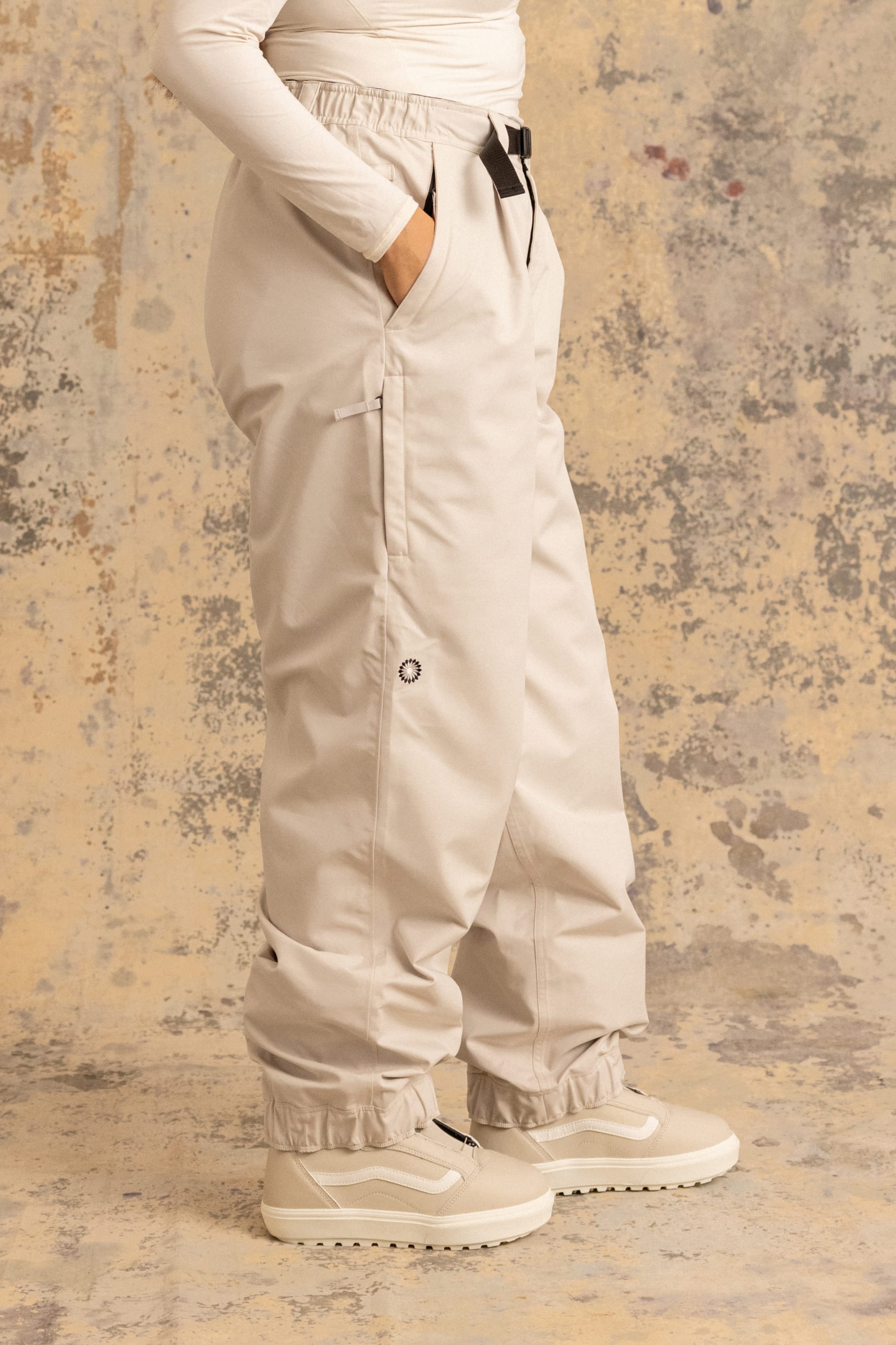 A person stands against a speckled background, wearing the 686 Women's Outline™ Shell Pant in light beige by 686, paired with a matching long-sleeve top and beige winter boots. Hands are in the pants pockets; only the lower body is visible.