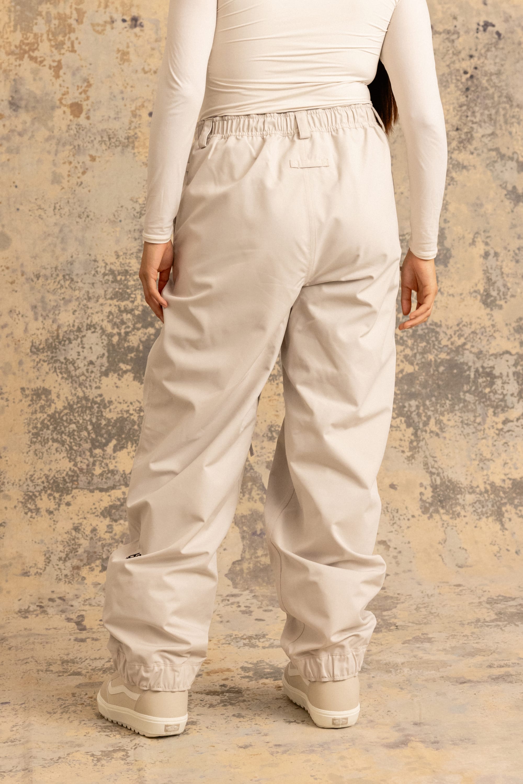 A person stands facing away, wearing the 686 Women's Outline™ Shell Pant by 686, paired with a fitted long-sleeve top and white shoes, set against a beige and gray textured background.