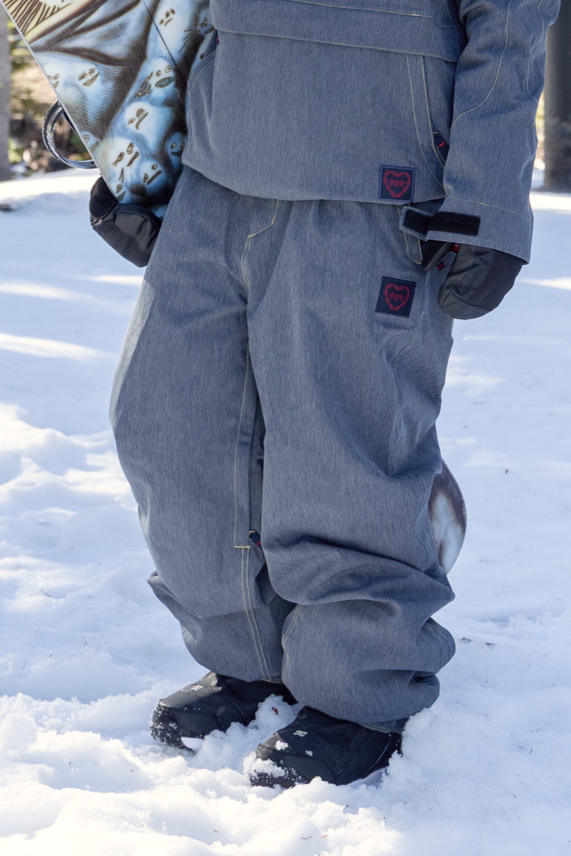 A person wearing the 686 Women’s Outline™ Shell Pant stands on snow, holding a snowboard, with only their lower body visible.