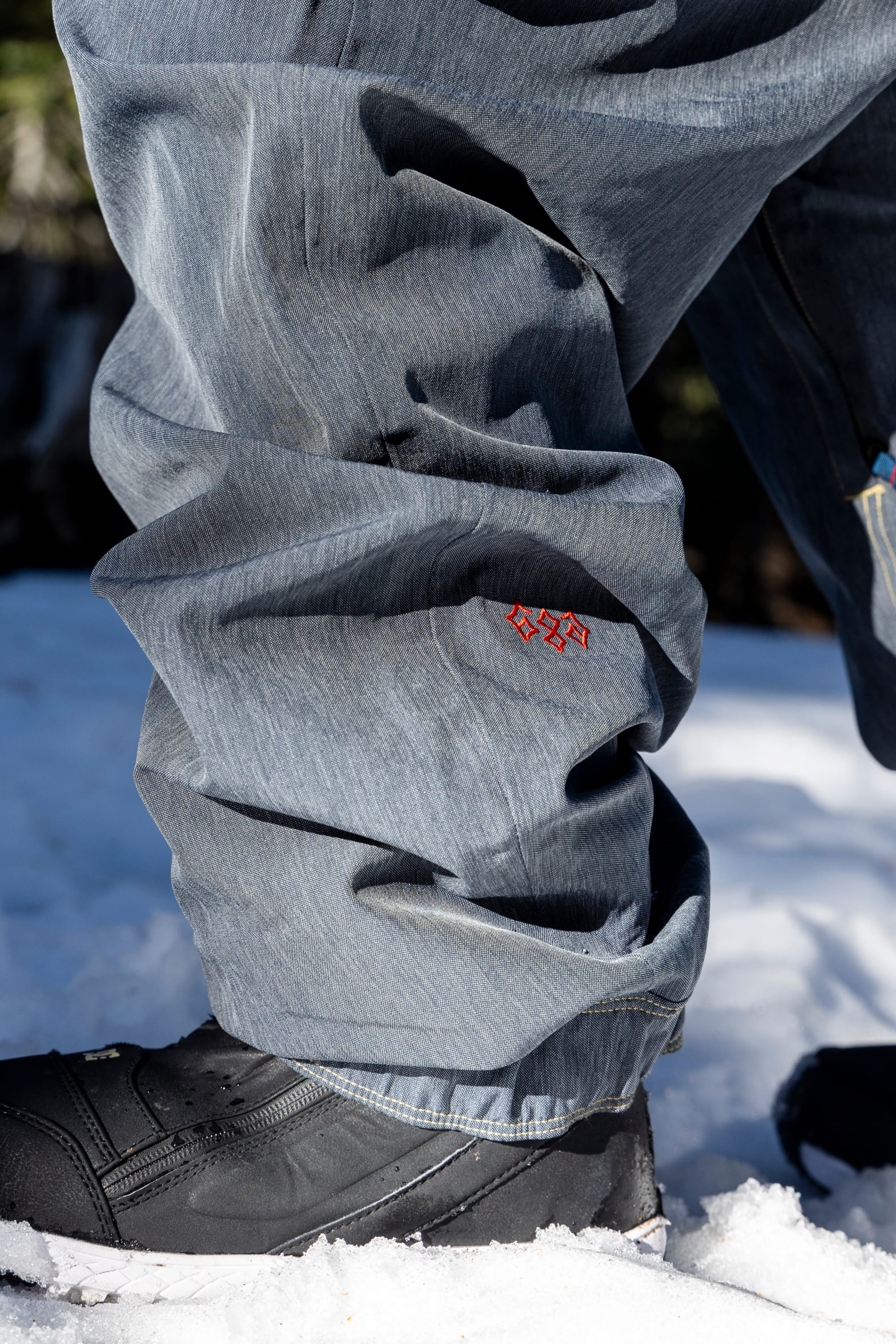 A close-up of a person standing on snow, wearing the 686 Women’s Outline™ Shell Pant in gray with a small red logo and black winter boots.