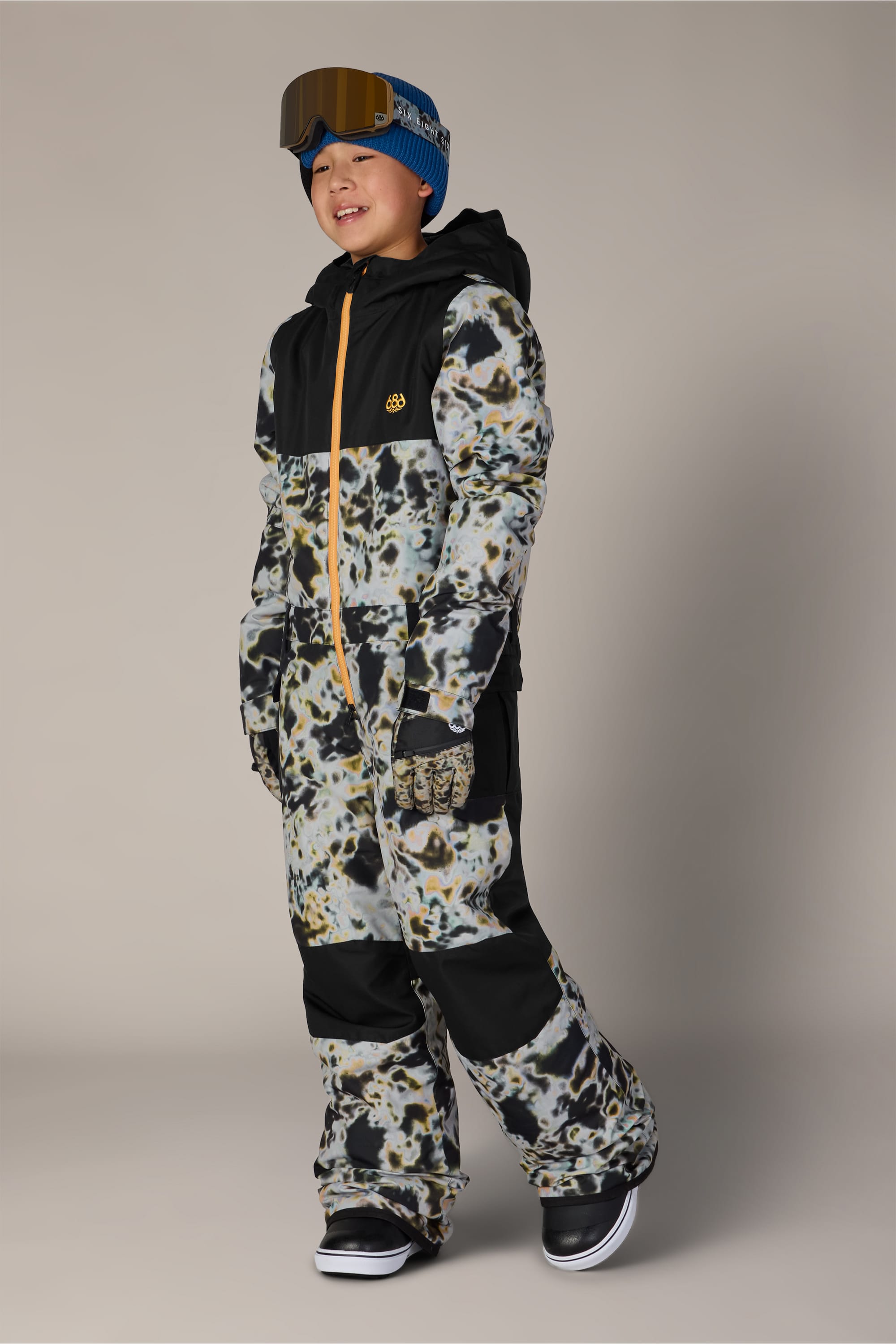 A child smiles against a plain background while wearing the 686 Boys’ Shazam Insulated One-Piece by 686, paired with a blue beanie and ski goggles.
