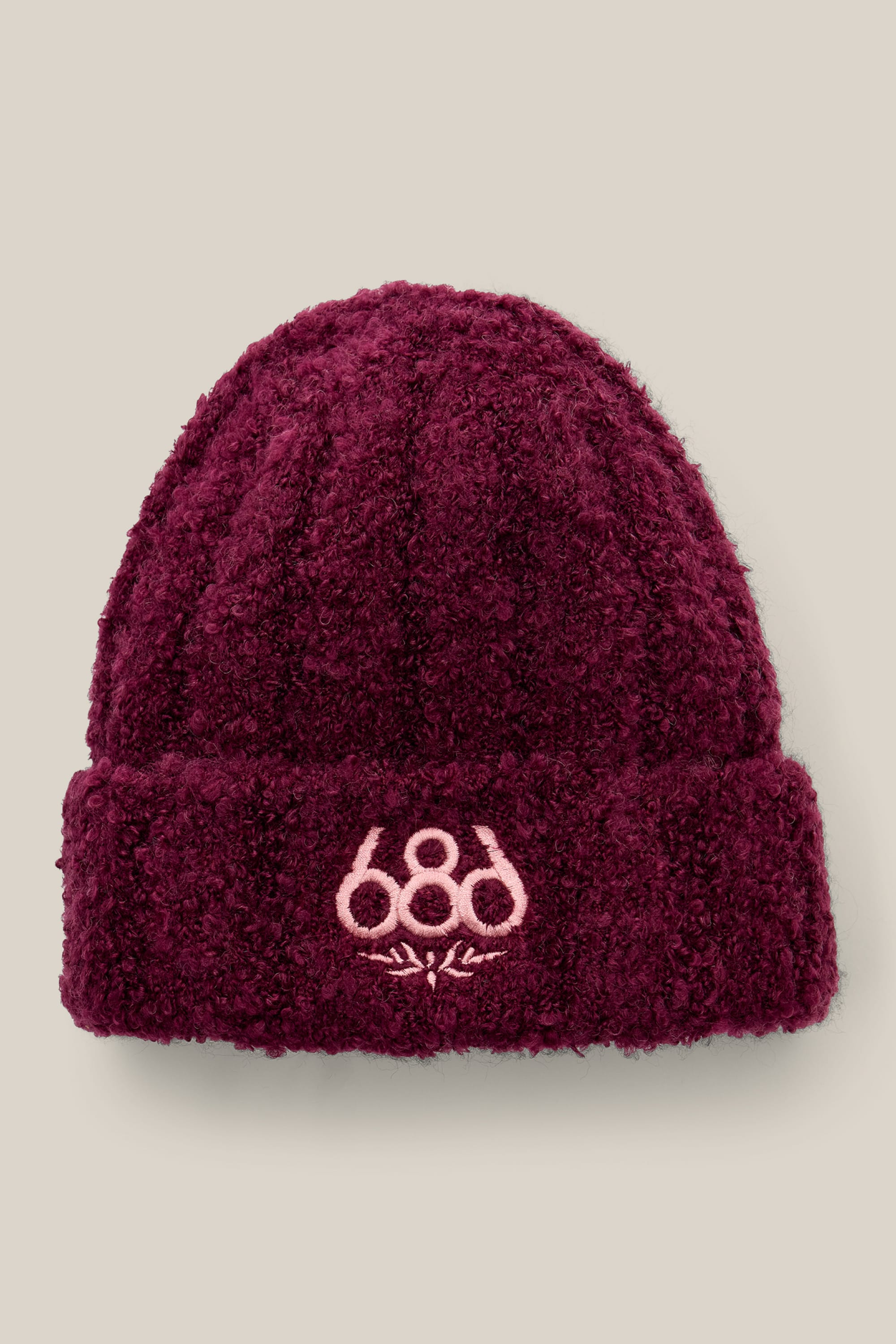 The 686 Women’s Fuzzy Beanie is a maroon knit hat with a folded brim, showcasing a light pink 686 logo and snowflake design on the front. The background features a solid light beige color.