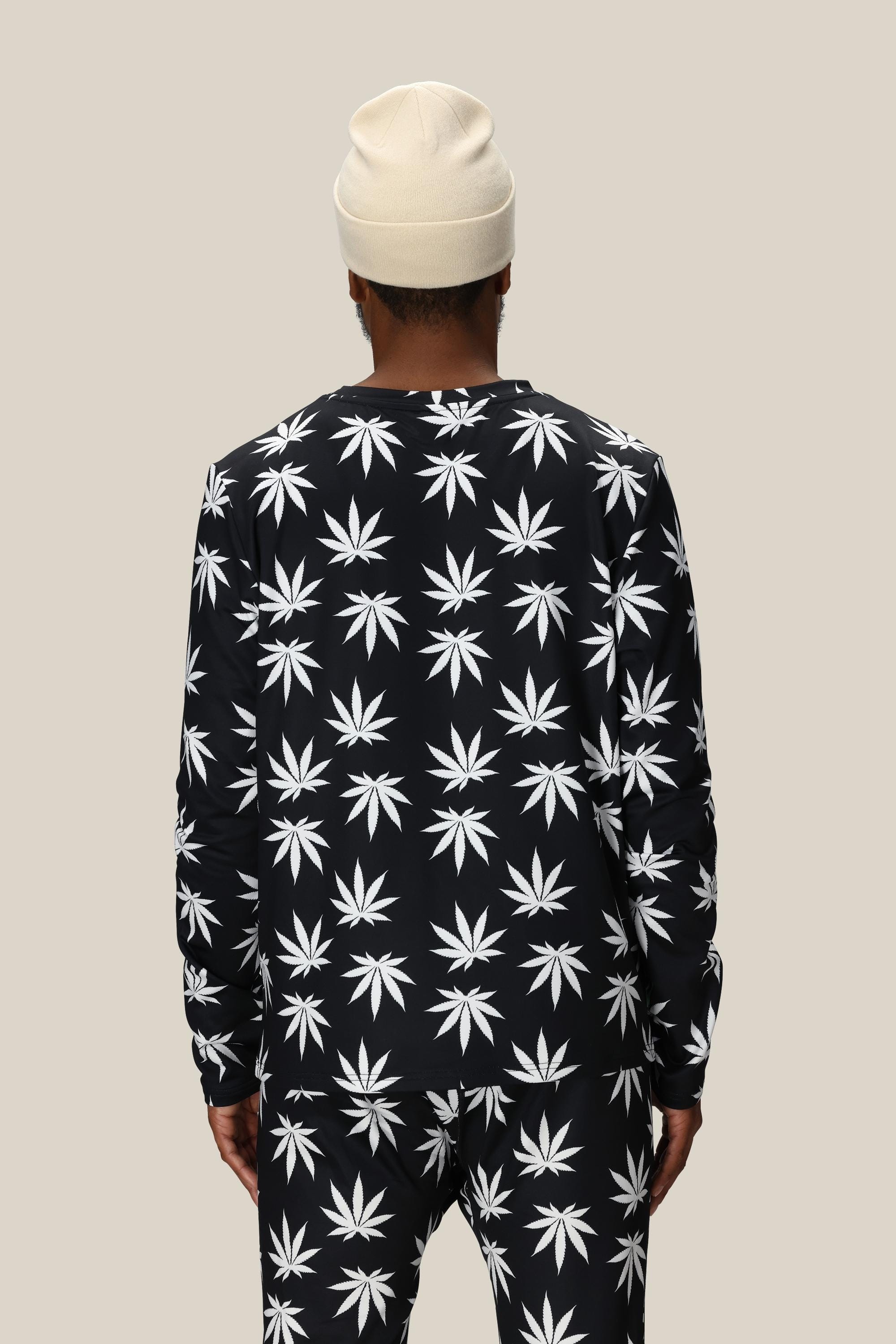 A person is seen from behind wearing the 686 Mens Plantlife Base Layer LS Top, featuring a white cannabis leaf pattern and paired with matching pants and a cream beanie, against a plain beige background.