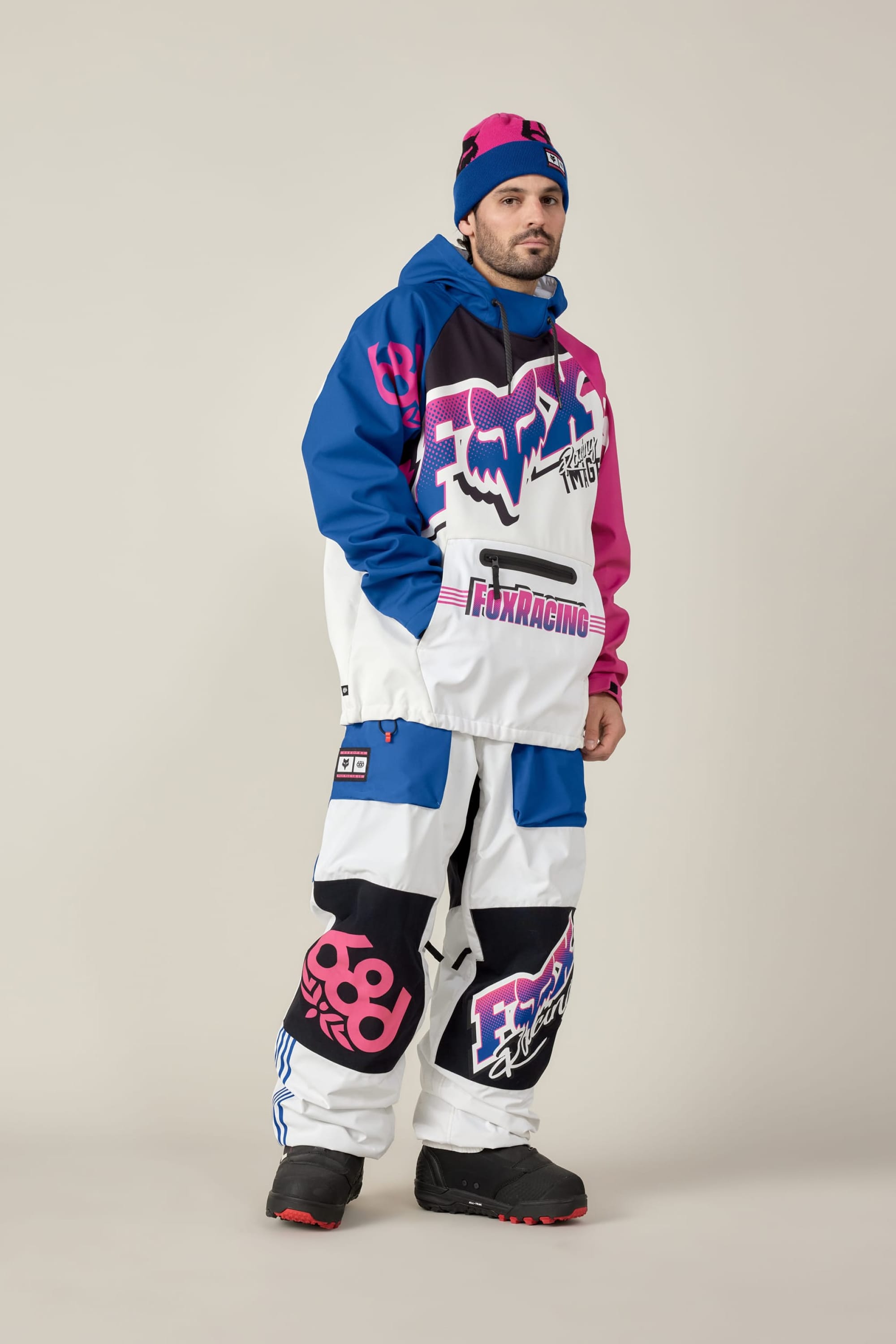 A man stands against a plain background wearing the 686 Men's Fox Racing Waterproof Hoody‚Äîblue, pink, and white with bold graphics‚Äîpaired with matching baggy pants, a blue-pink beanie, and black snow boots.