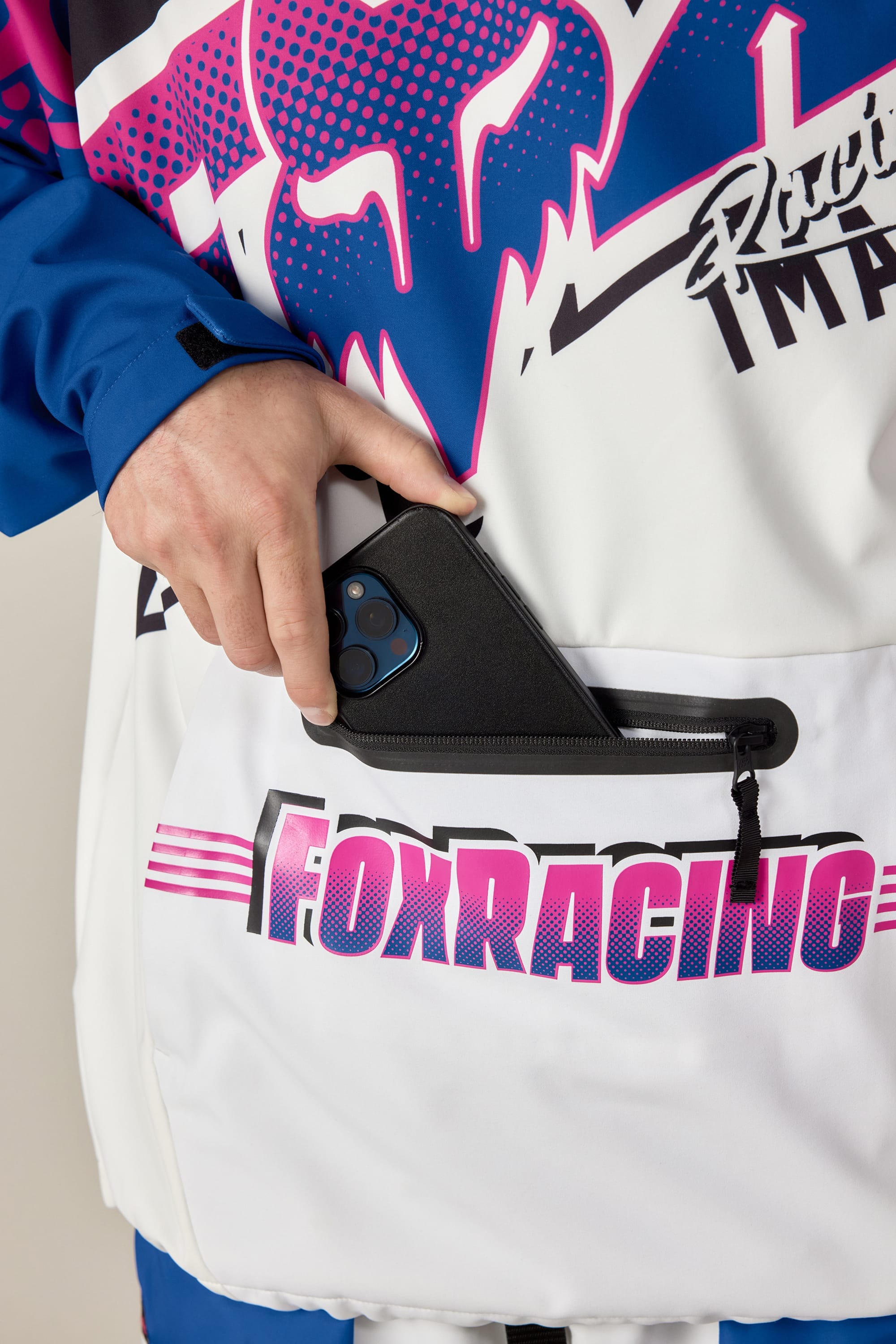 A person wearing a 686 Men's Fox Racing Waterproof Hoody with bold pink and blue graphics puts a black smartphone into the jacket‚Äôs front zippered white pocket.